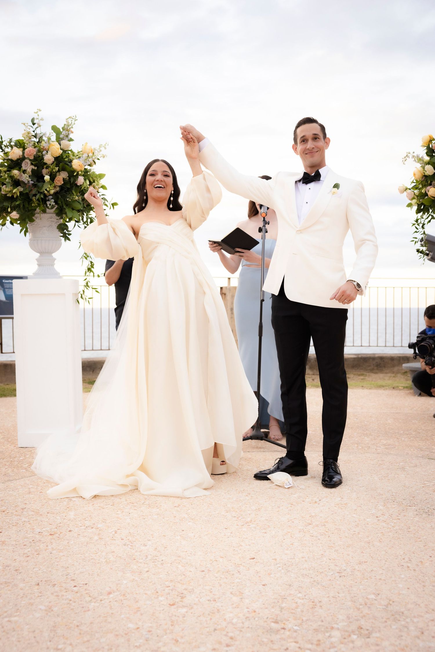 Wedding couple poses on stone terrace with floral arrangements and formal attire.