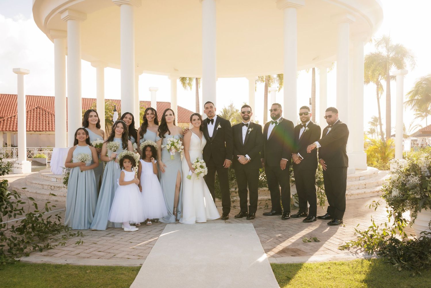 A wedding party poses together under a white gazebo surrounded by palm trees and landscaping on a sunny day.