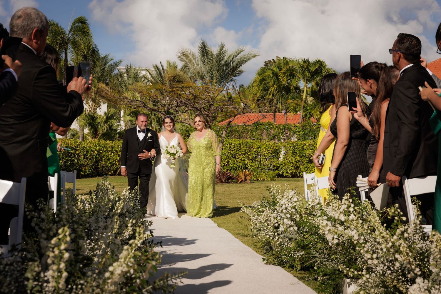 Guests gather at an outdoor wedding ceremony set in a lush tropical garden with palm trees and blooming flowers.