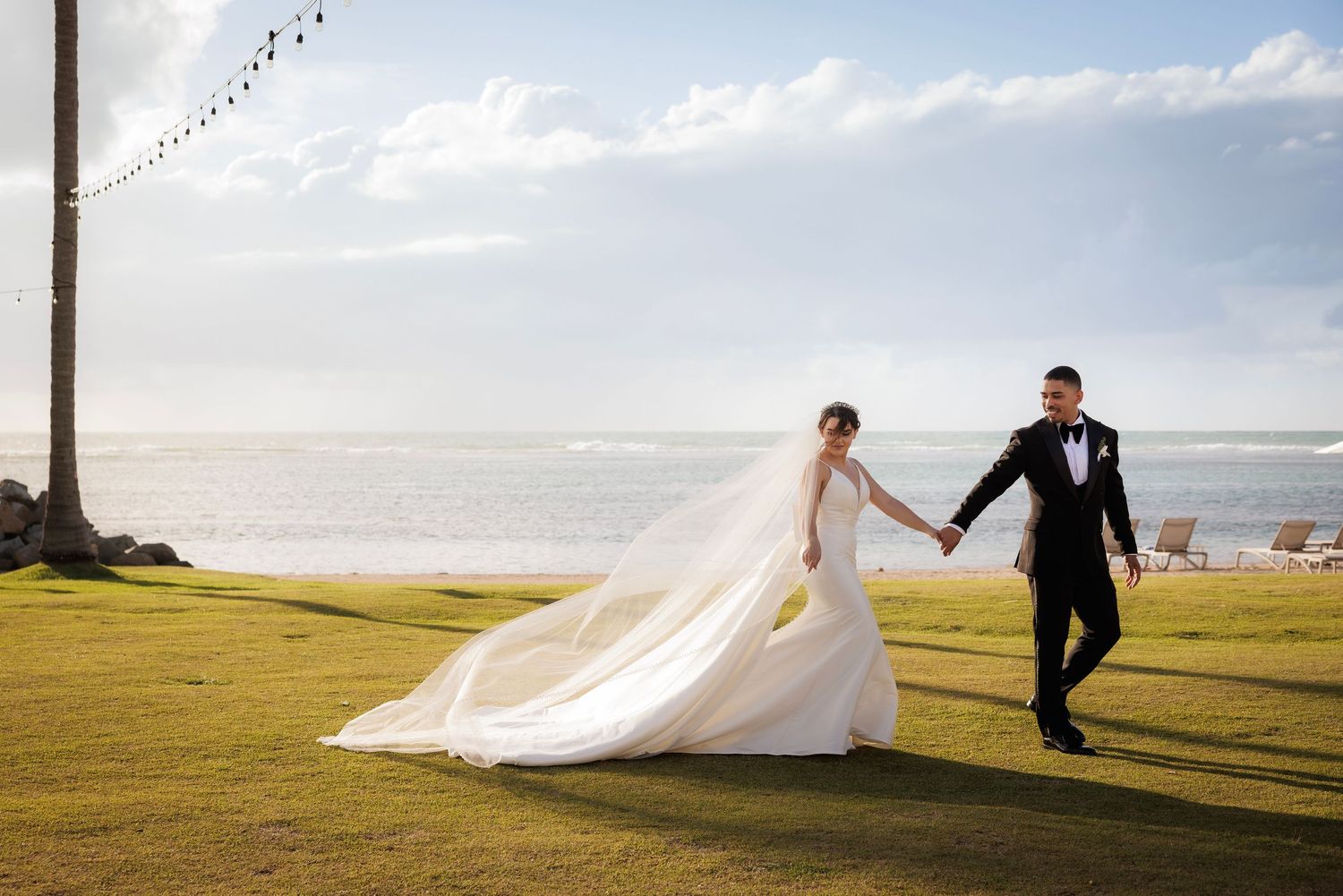 Two people in wedding attire run playfully along a scenic coastal cliff with a flowing white dress in the wind.