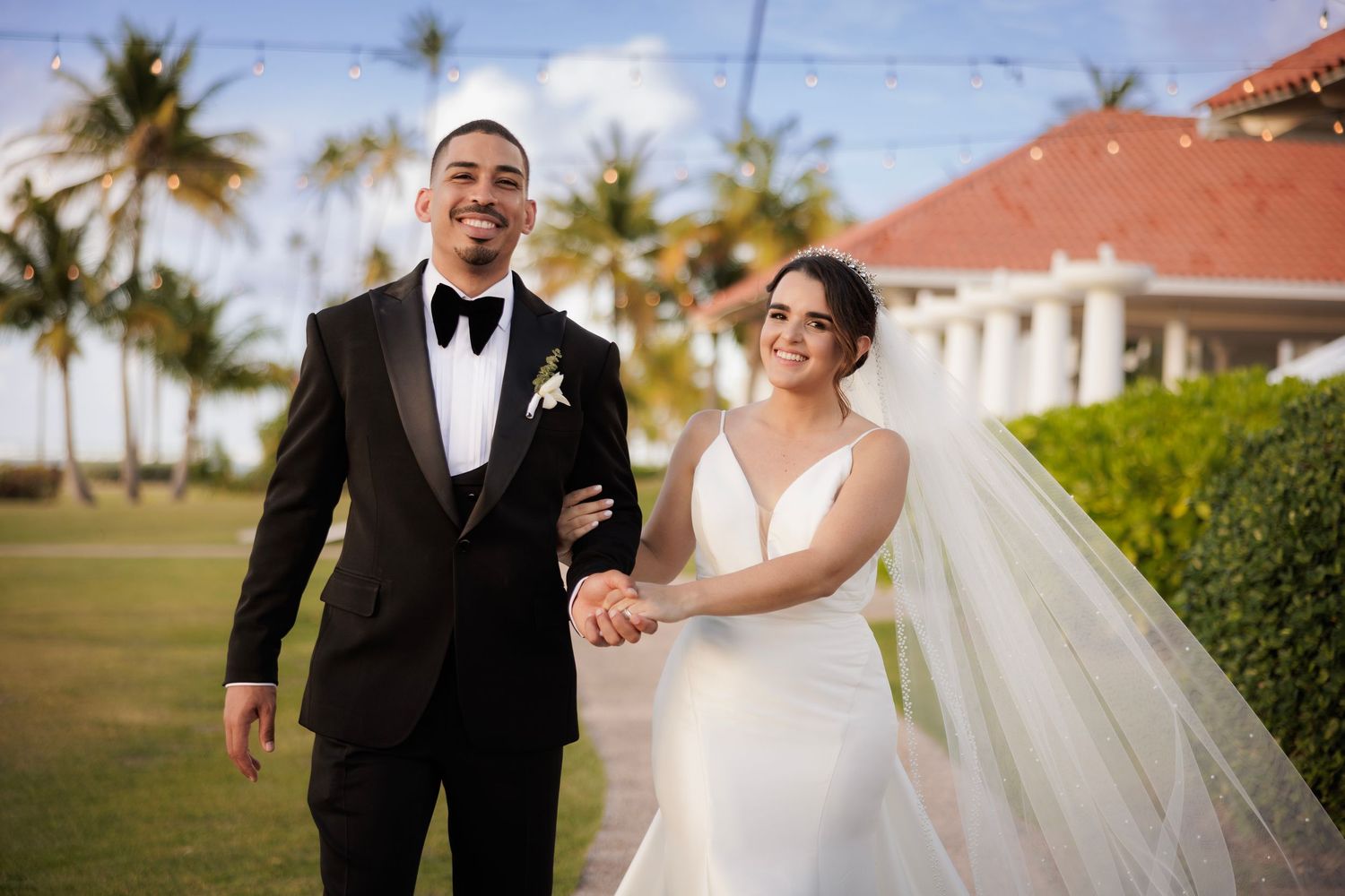 A couple in wedding attire holds hands while walking and smiling together on a sunny outdoor path lined with palm trees.