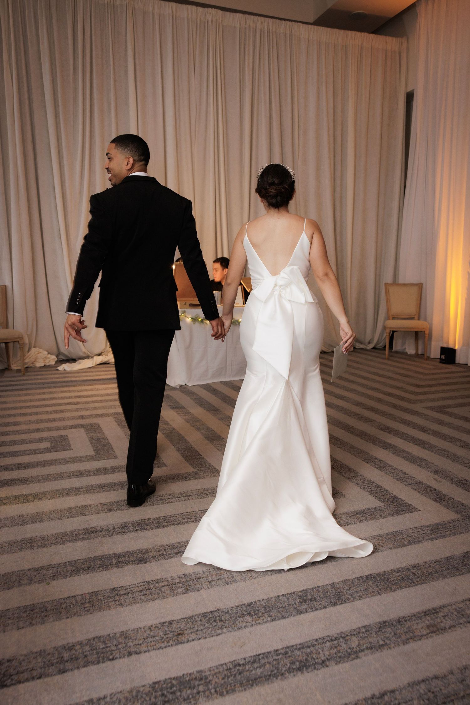A couple in formal wedding attire walks hand in hand across a chevron patterned carpet in an elegant reception venue.