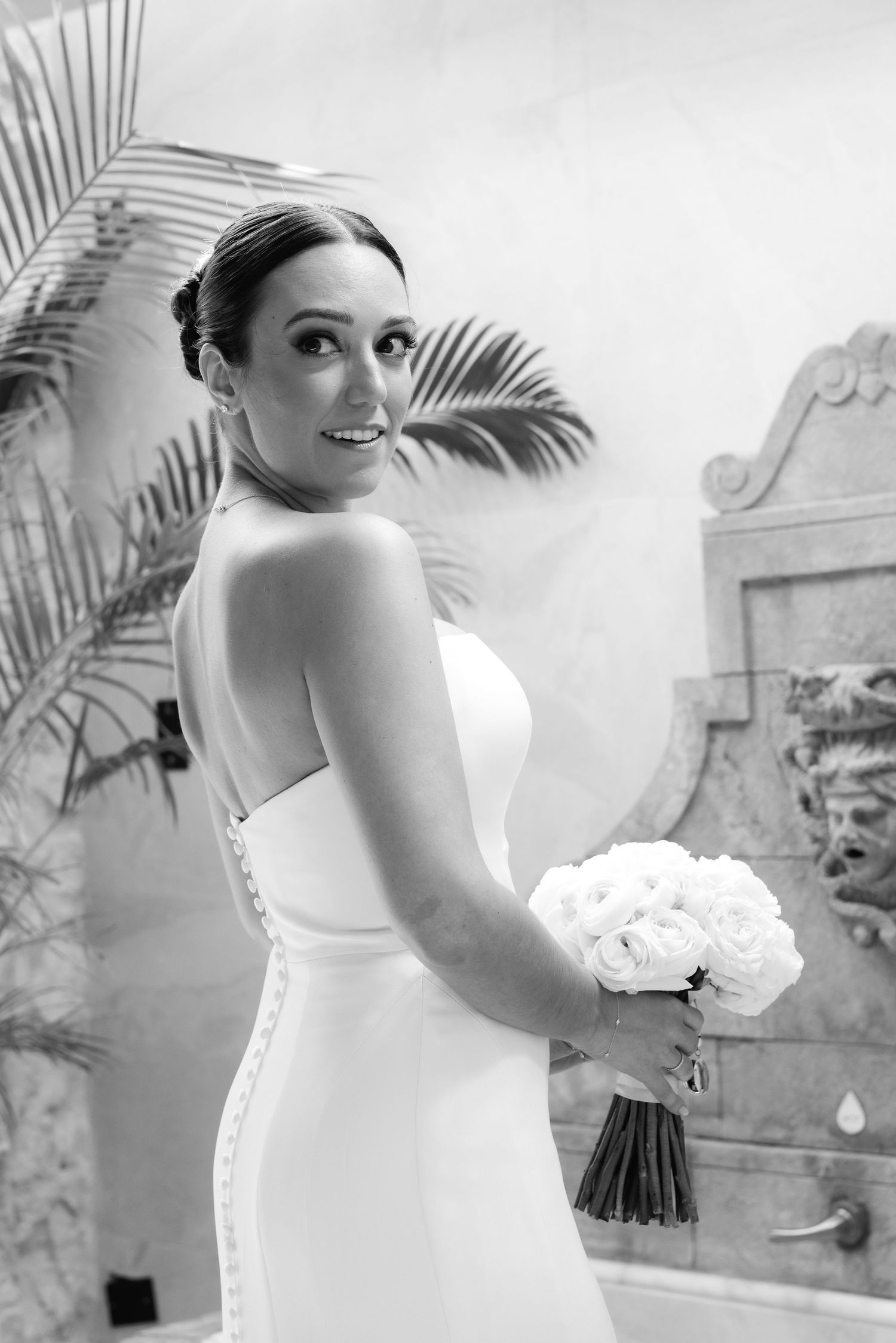A bride in a strapless white wedding gown holds a bouquet while posing against a tropical garden background.