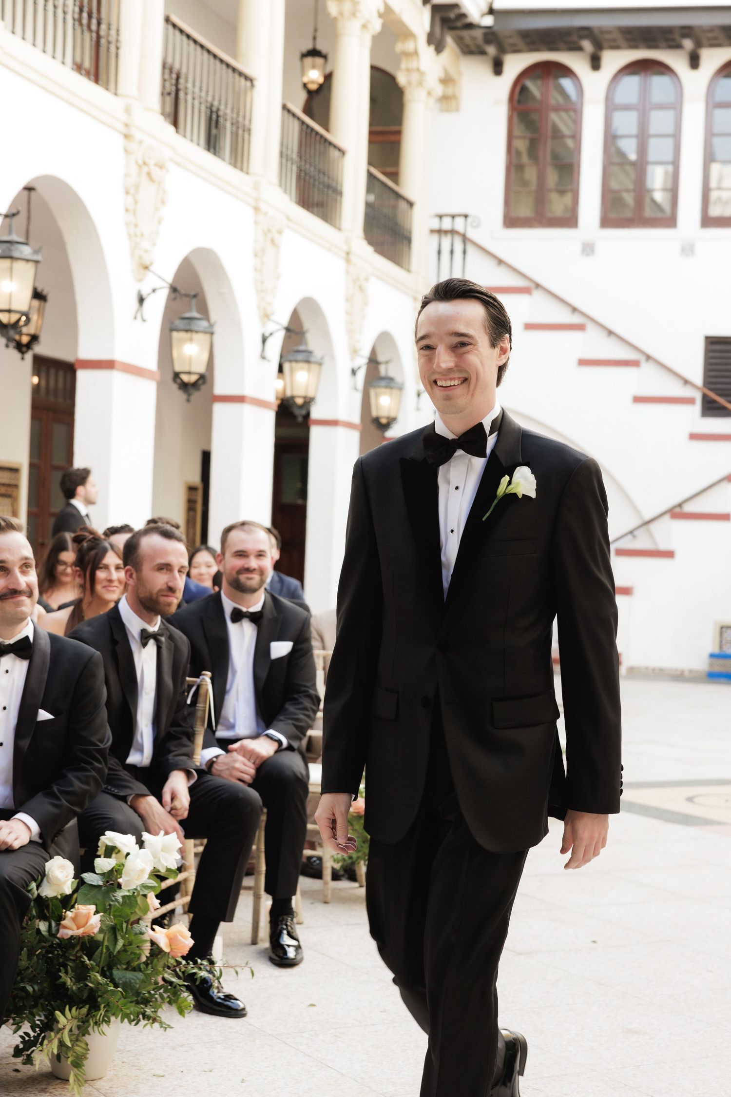 A wedding party smiles and shares candid moments in a bright courtyard with arched architectural details.