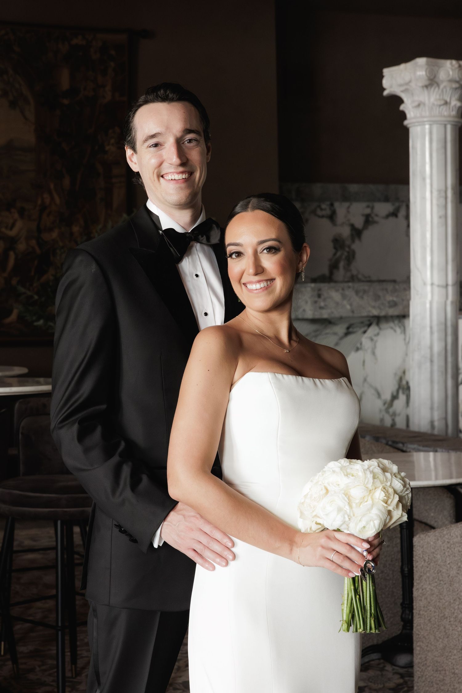 Wedding couple share intimate moments together in formal attire under marble columns at elegant venue.