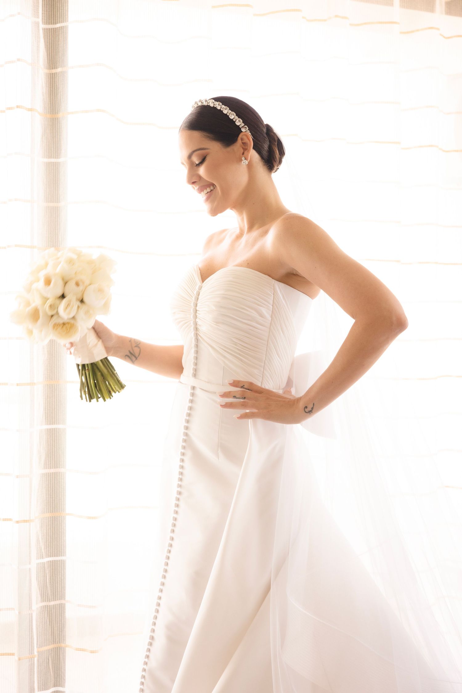 A radiant bride in a strapless white wedding gown holds a white bouquet while posing by a window.