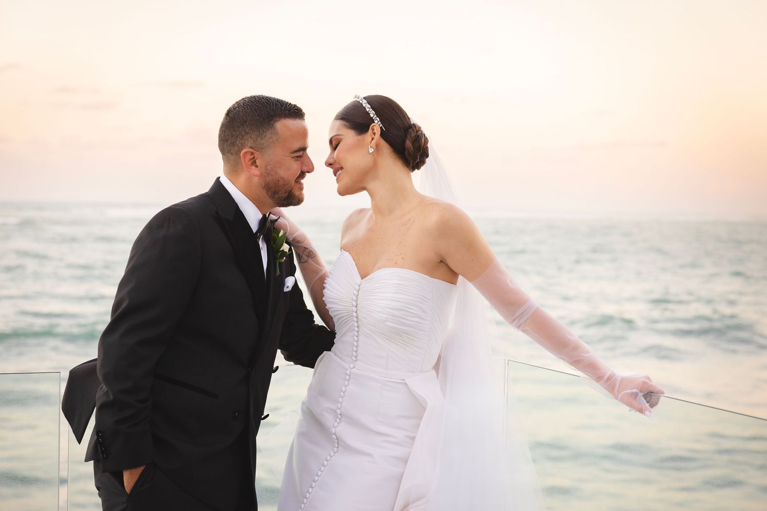 A romantic couple in wedding attire embrace at sunset on a beach with the ocean in the background.