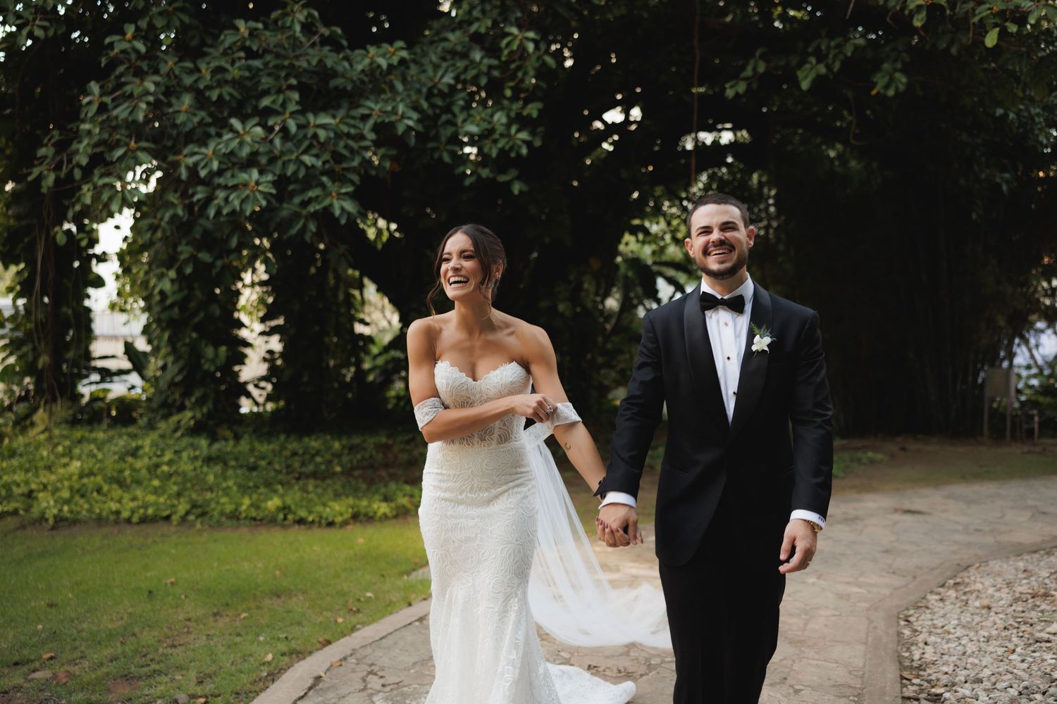 Happy couple walks on garden path during outdoor wedding celebration at sunset.