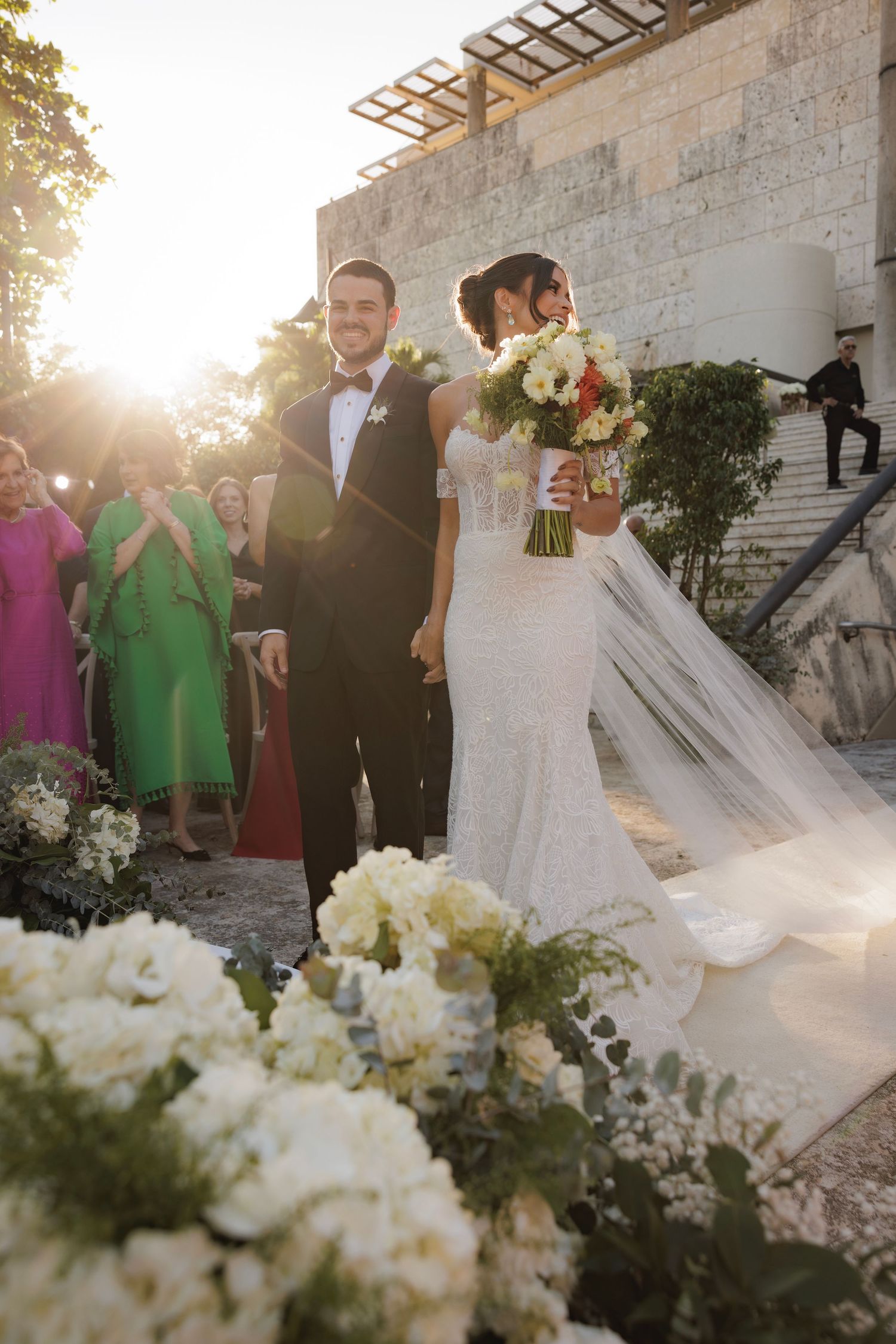 A sun-drenched wedding ceremony featuring white floral arrangements and guests in colorful attire.