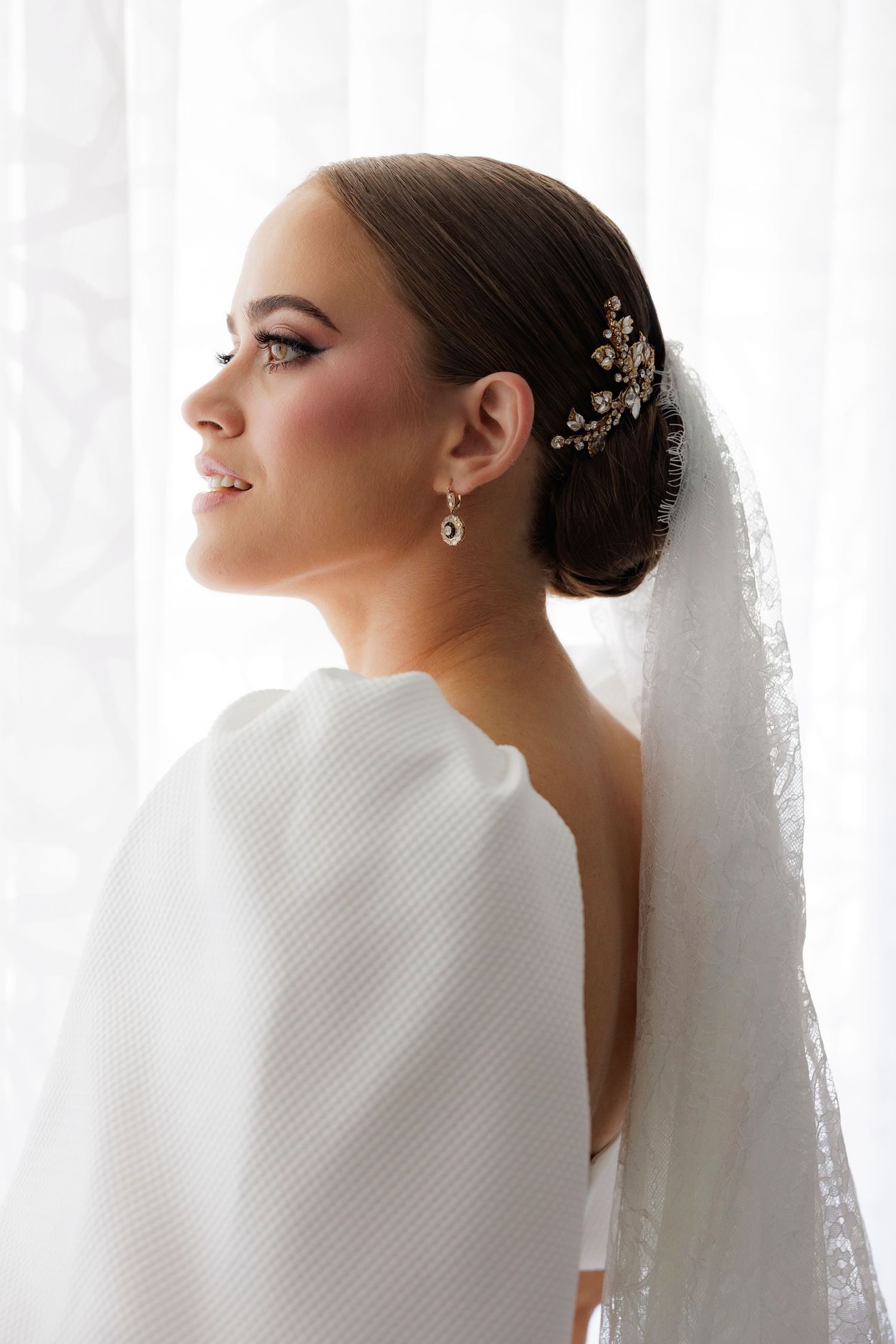 Elegant bridal portrait in profile showing delicate veil, floral hair accessory and pearl drop earring against white background.