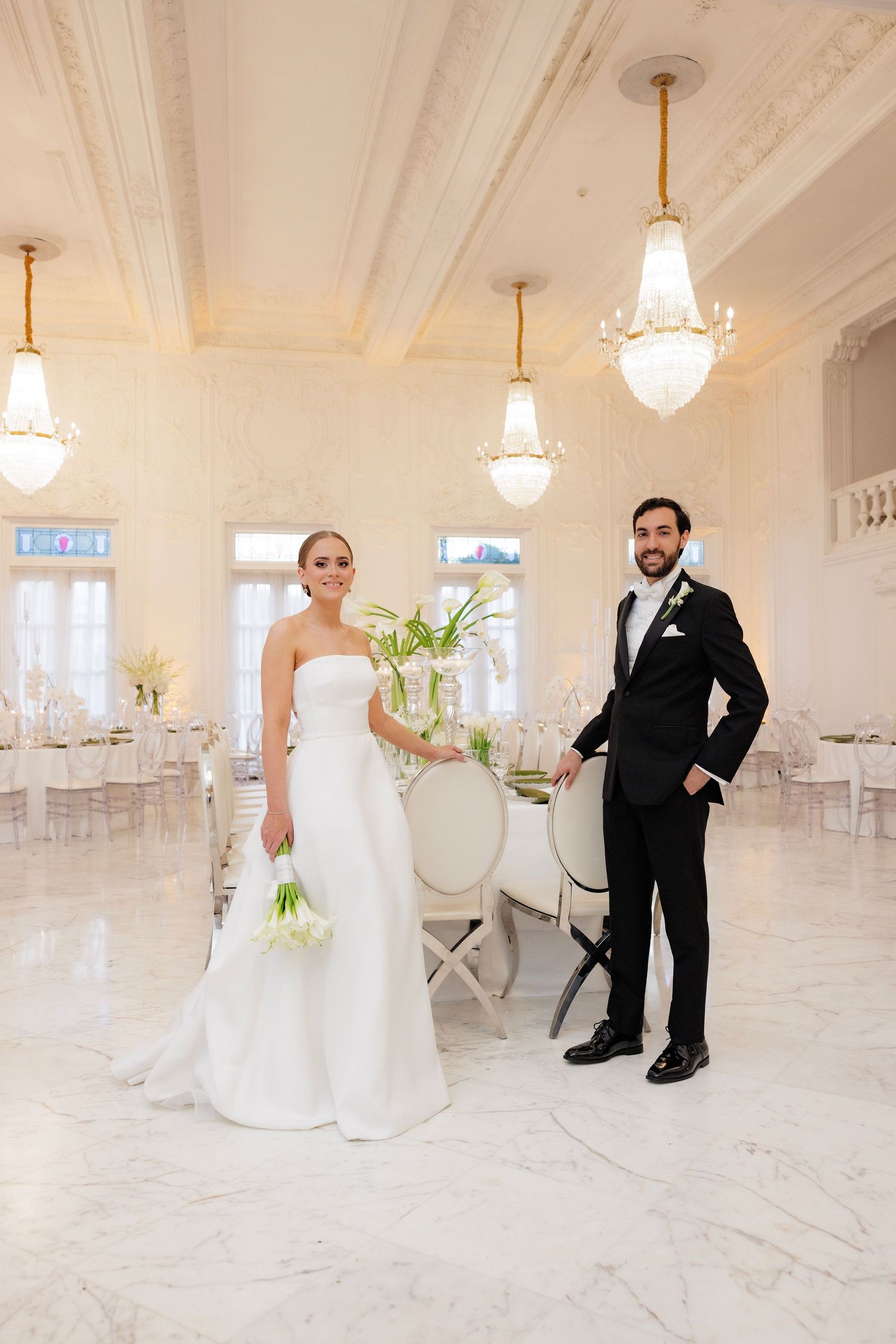 A wedding couple stands in an elegant white ballroom with crystal chandeliers and marble floors.