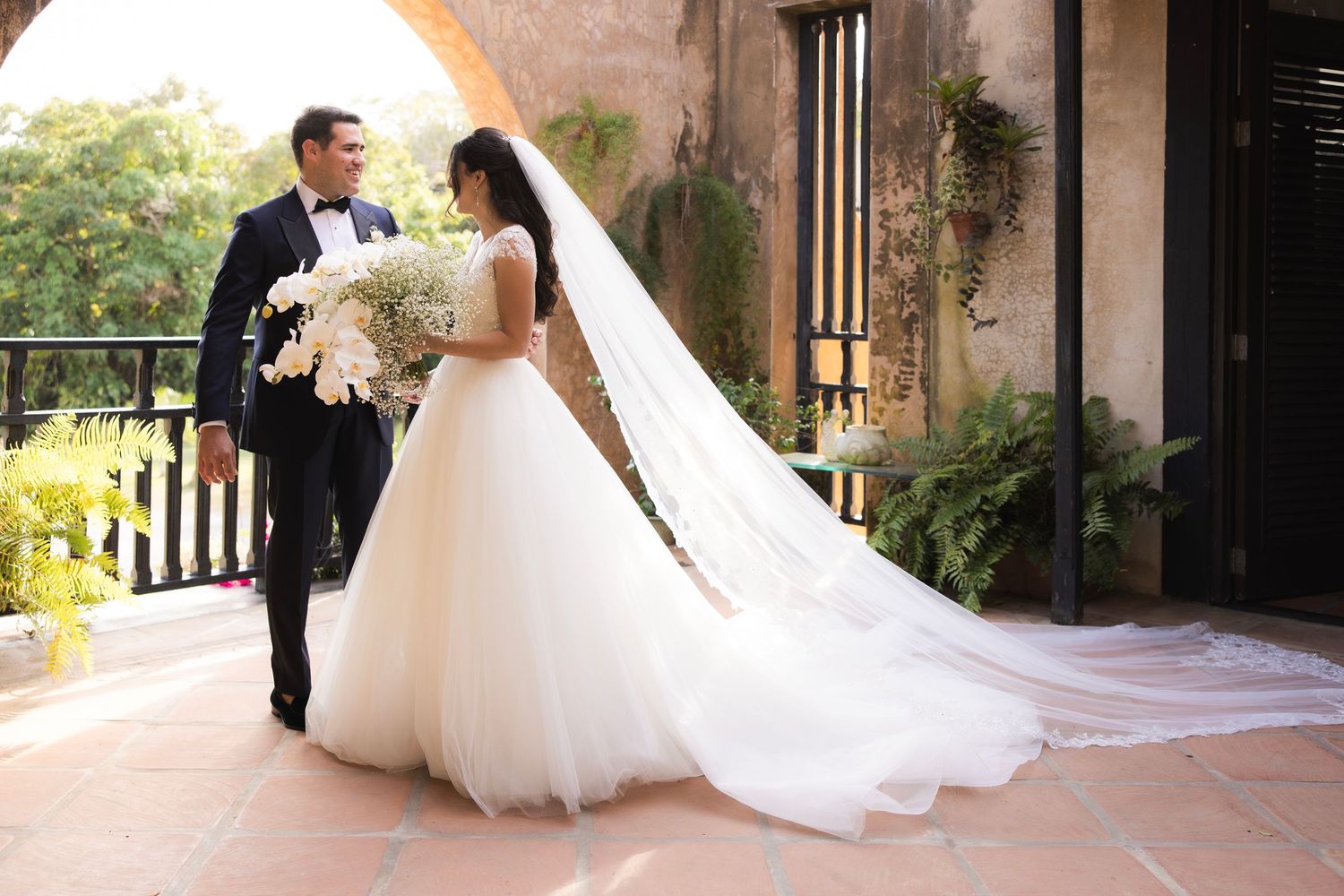 Bride and groom walk along a sunlit stone courtyard with flowing white wedding dress.