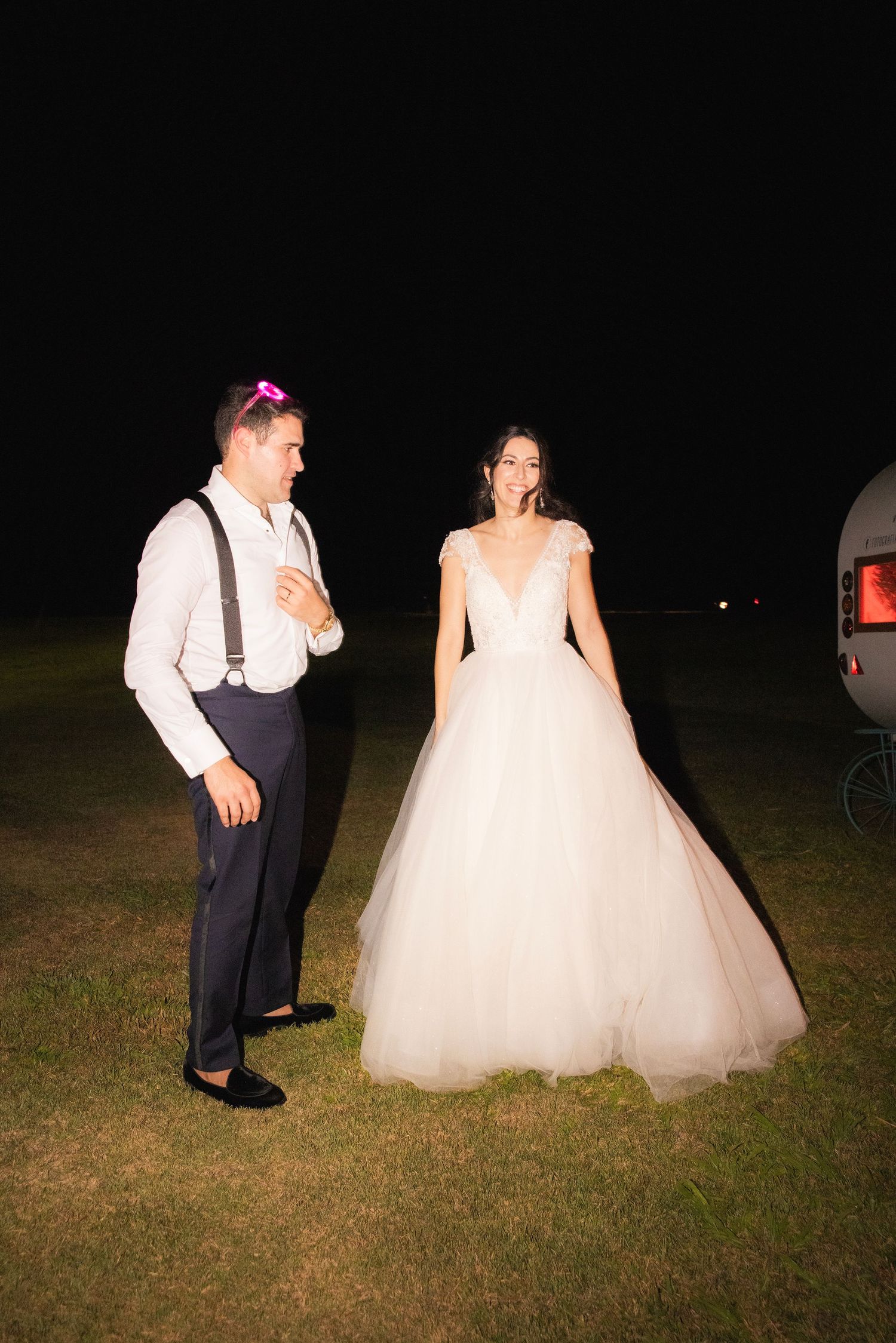 A couple in wedding attire poses outdoors at night on a dark grass field.