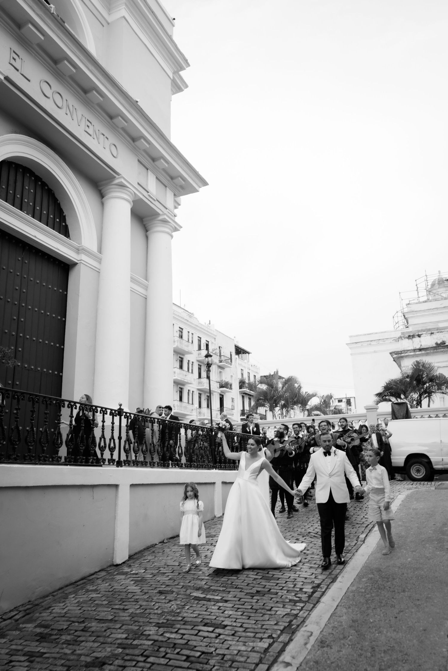 A wedding party walks along a cobblestone street outside a historic white church building.
