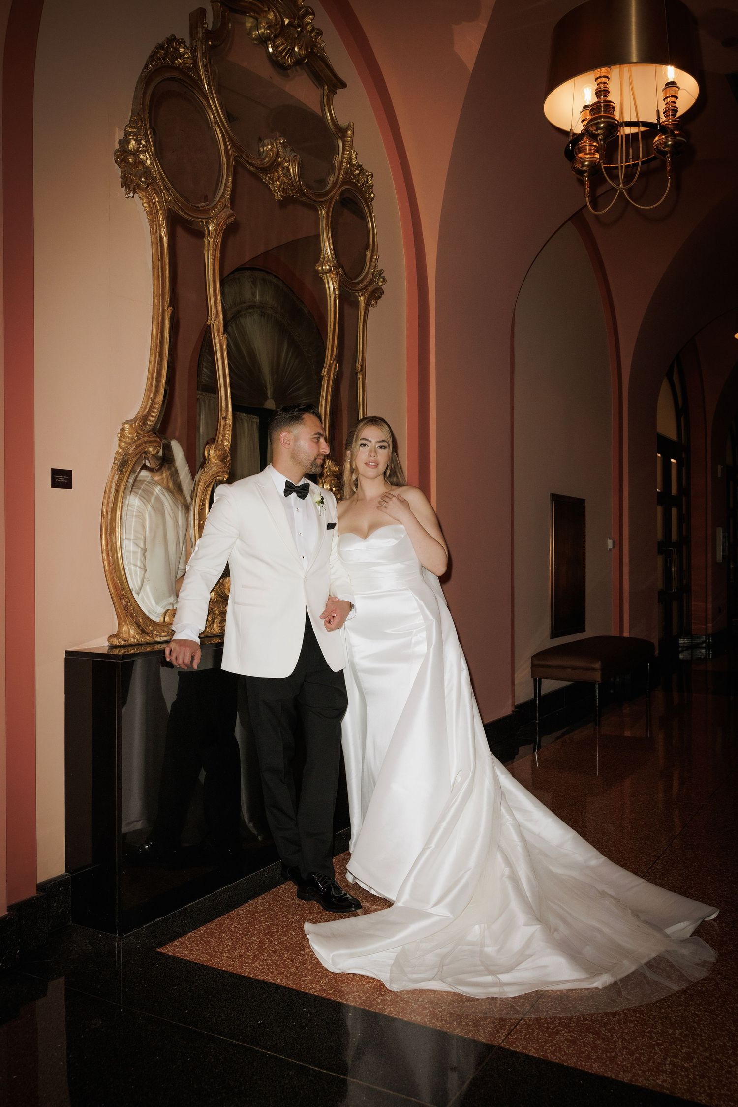A couple poses in formal wedding attire next to an ornate gold mirror in an elegant hallway with pink walls.