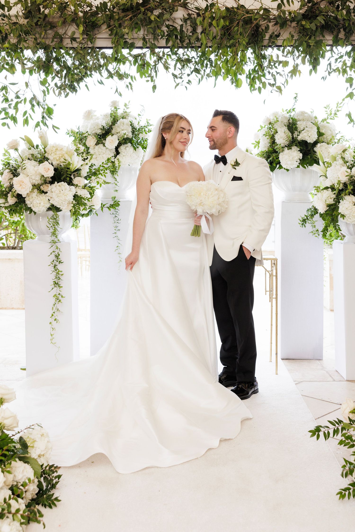 Romantic wedding portraits under a white pergola adorned with lush florals and hanging greenery.