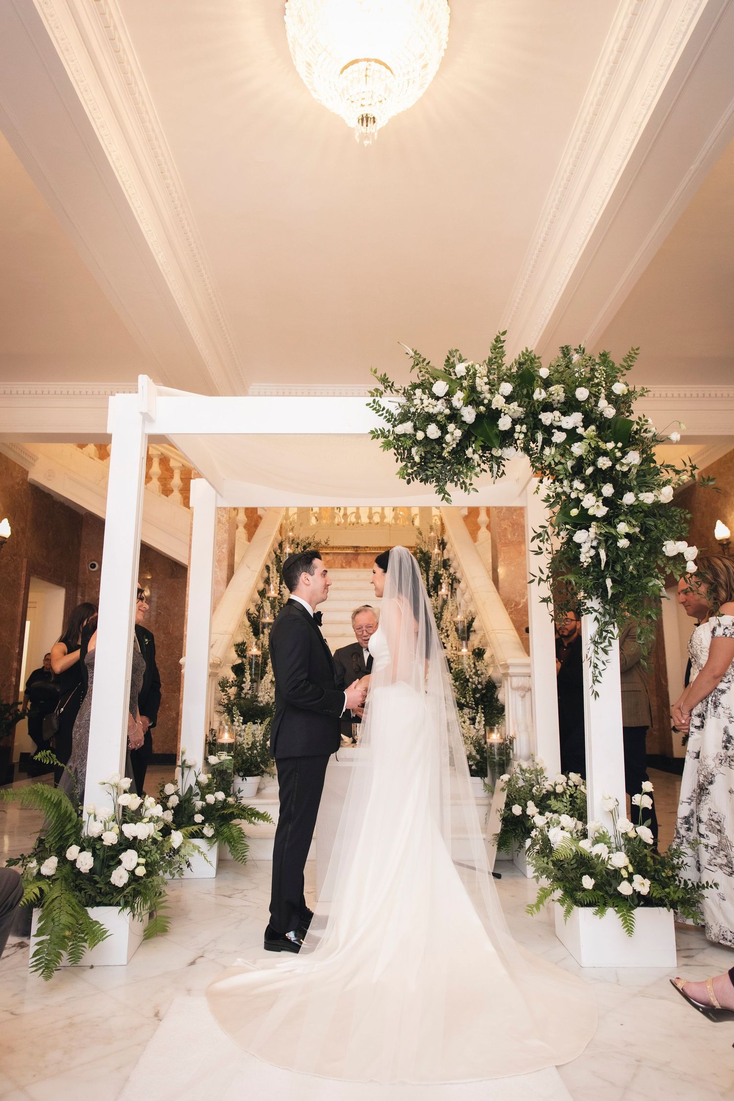 Elegant indoor wedding ceremony with white floral arrangements and decorative columns in a grand venue.