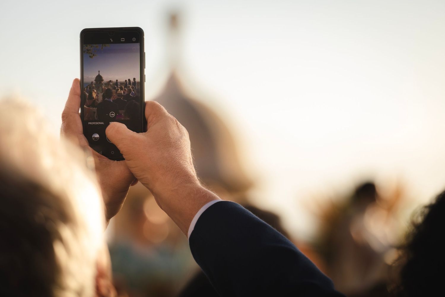 Silhouetted crowd holds up glowing mobile phones during outdoor concert or event at sunset or sunrise.