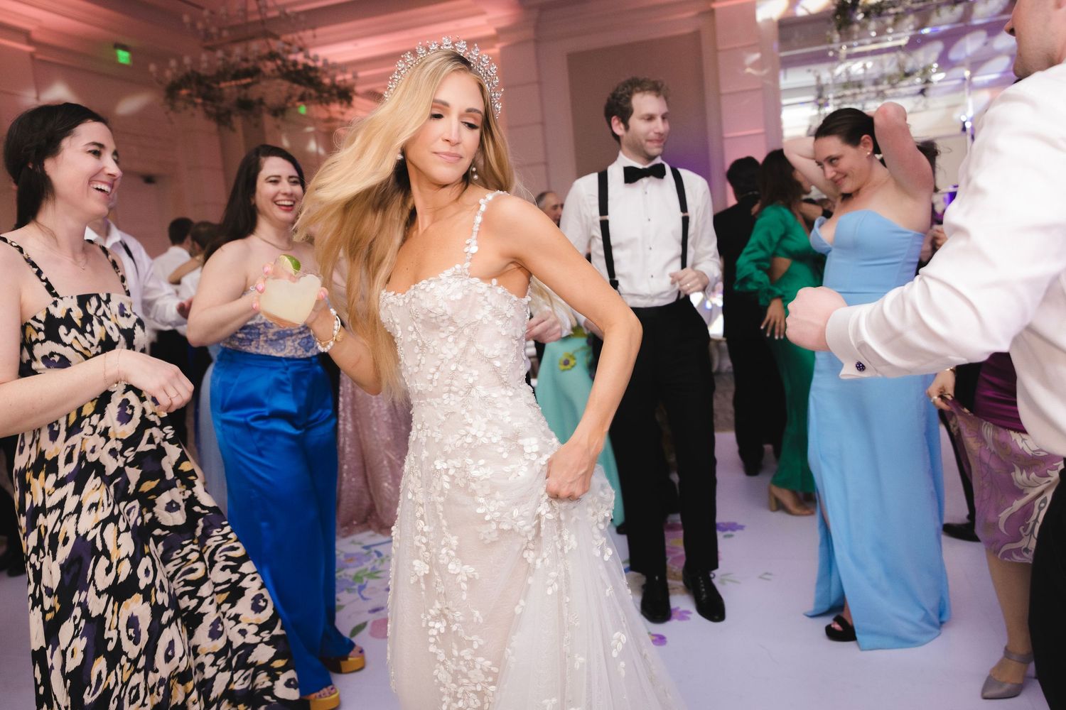 Party guests dance and celebrate at an elegant wedding reception with pink lighting in a grand ballroom.