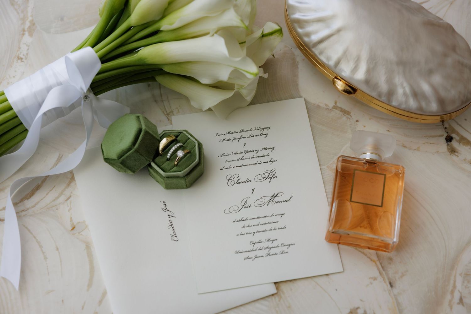 Wedding invitation displayed with white calla lily bouquet, green ring box and perfume bottle on white silk fabric.