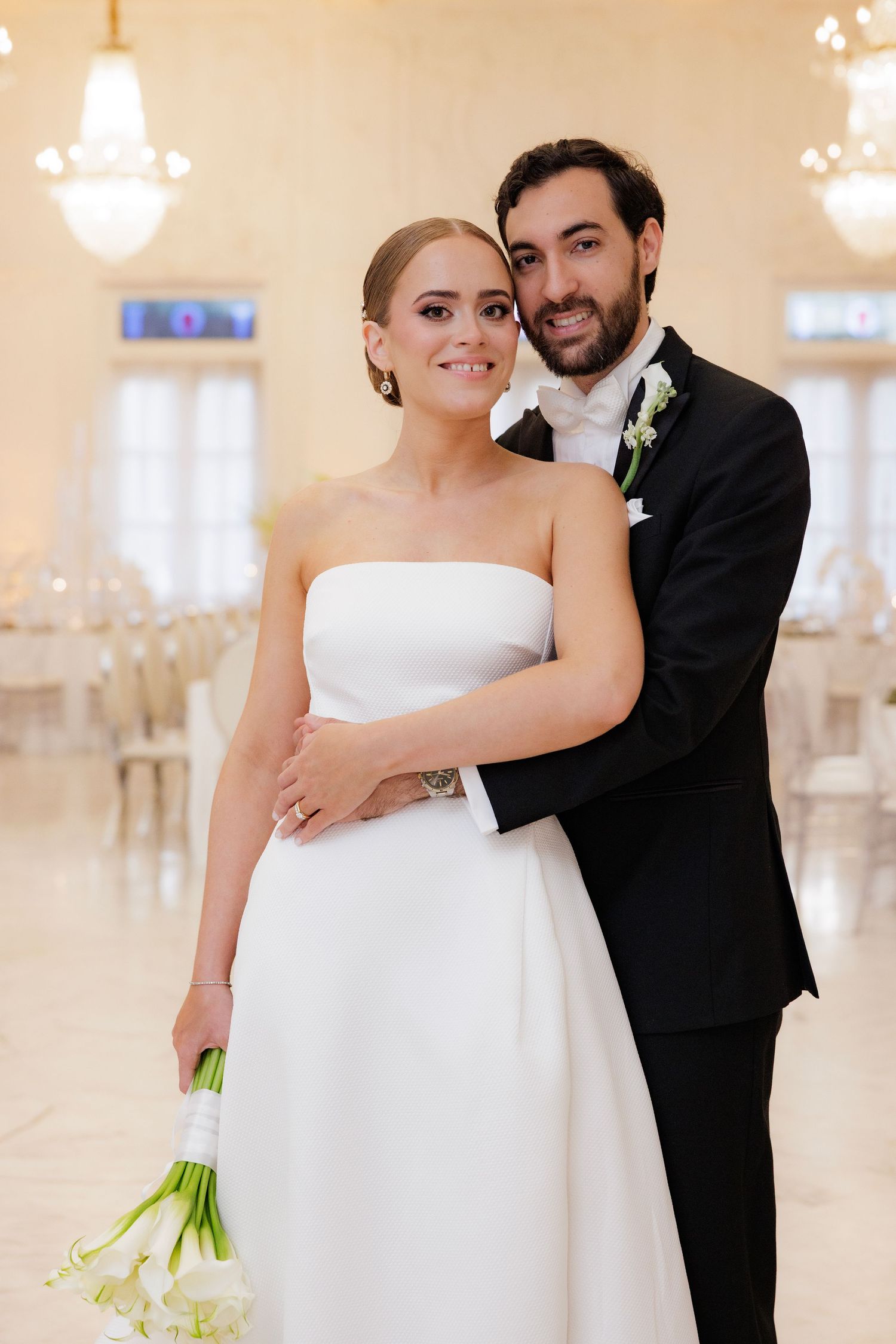 A couple in formal wedding attire poses together in an elegant ballroom with chandeliers.