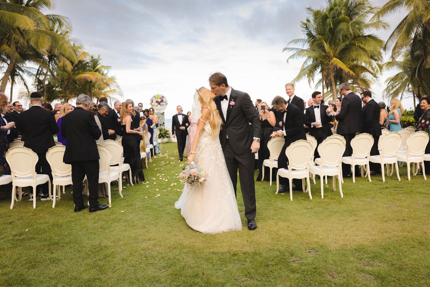 A wedding ceremony takes place on a lush green lawn with palm trees and white chairs under a cloudy tropical sky.