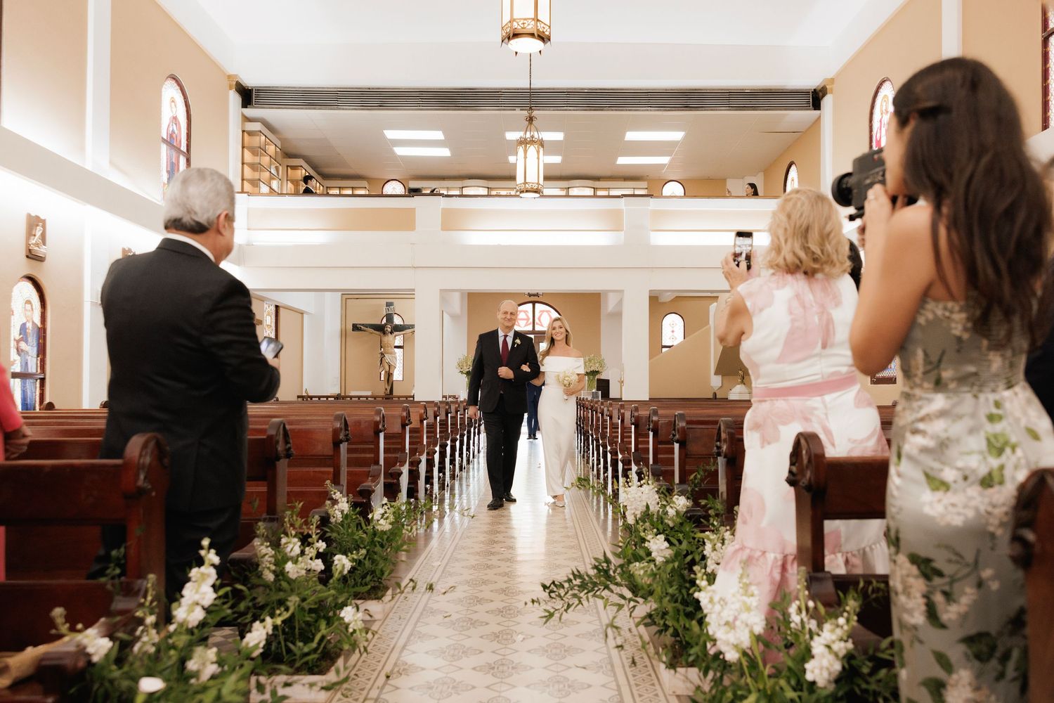 A church aisle decorated with elegant floral arrangements along the wooden pews for a wedding ceremony.