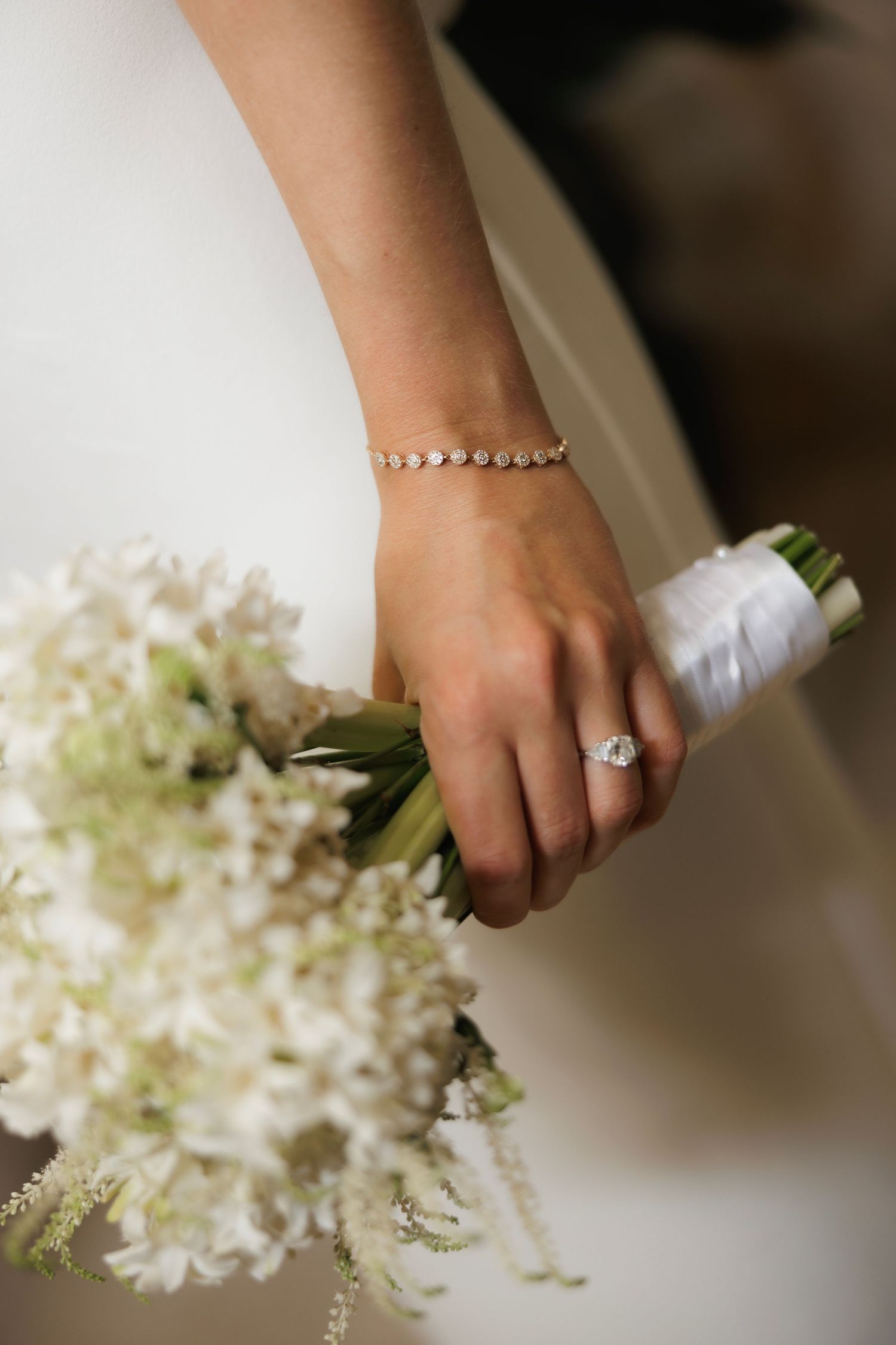 A bride's hand wearing an elegant diamond ring and pearl bracelet while holding a white floral bouquet.