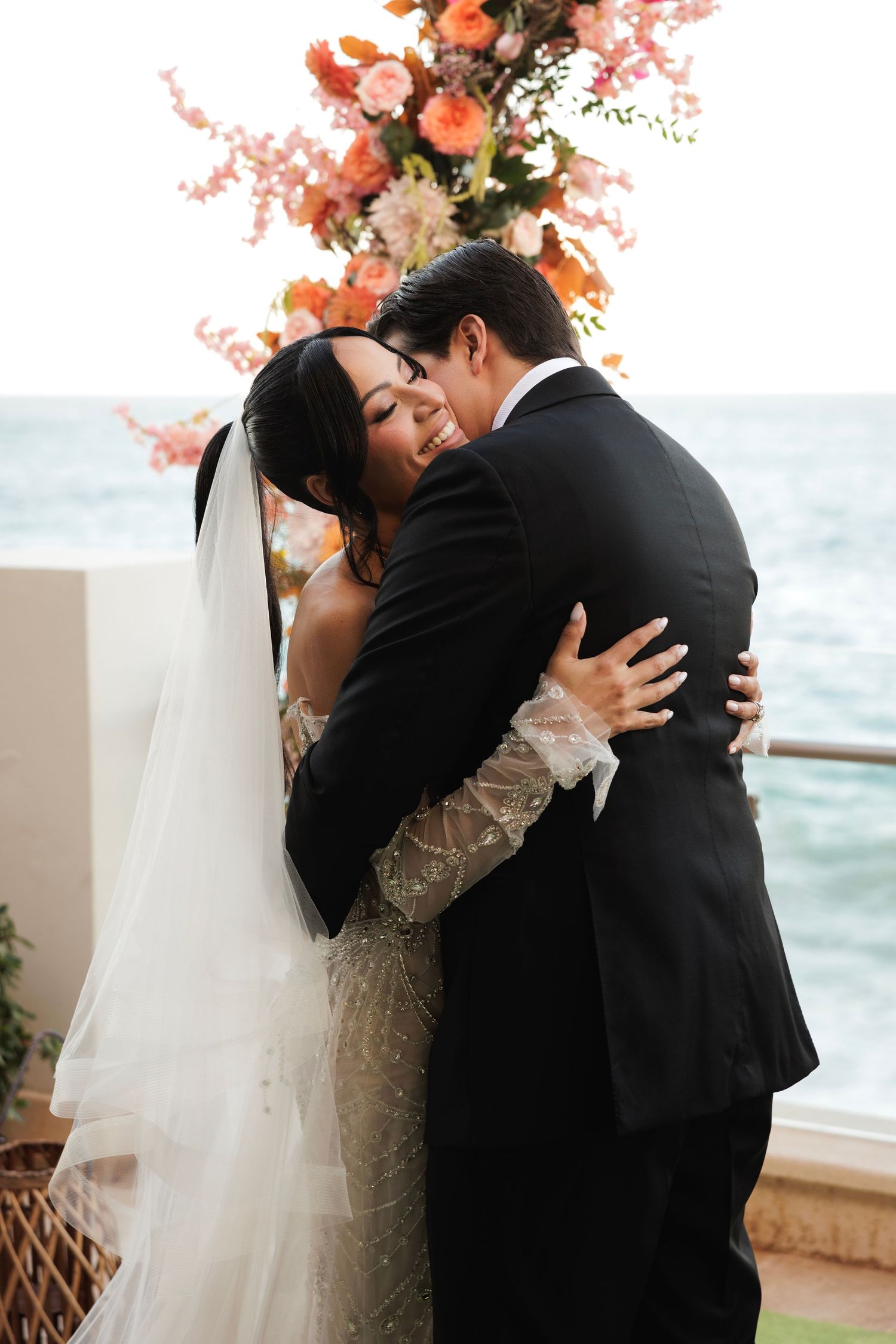 A romantic wedding couple shares their first kiss as newlyweds by the ocean with coral floral decorations.