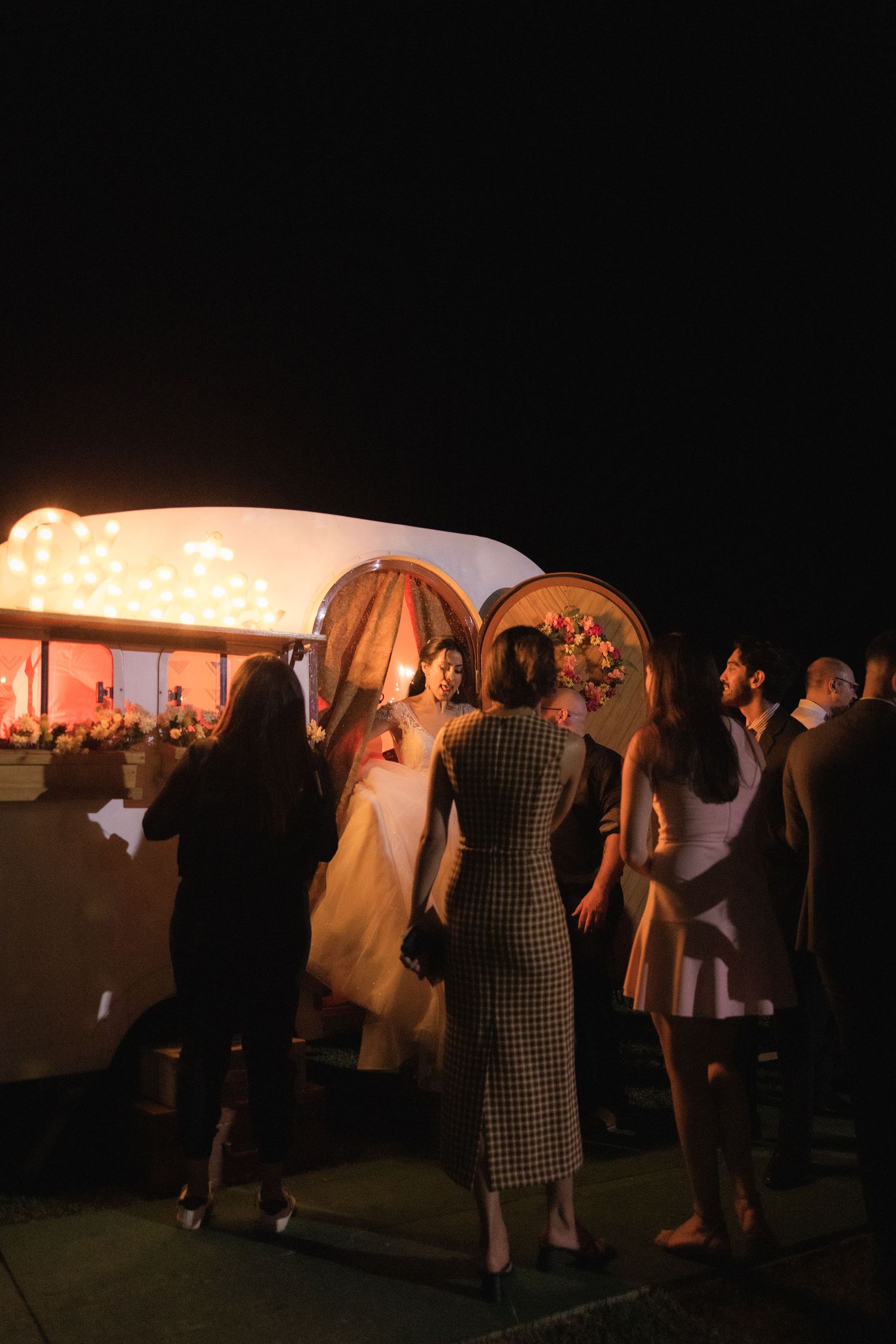 Wedding guests dancing at an outdoor evening reception with ambient lighting and tent structures visible.