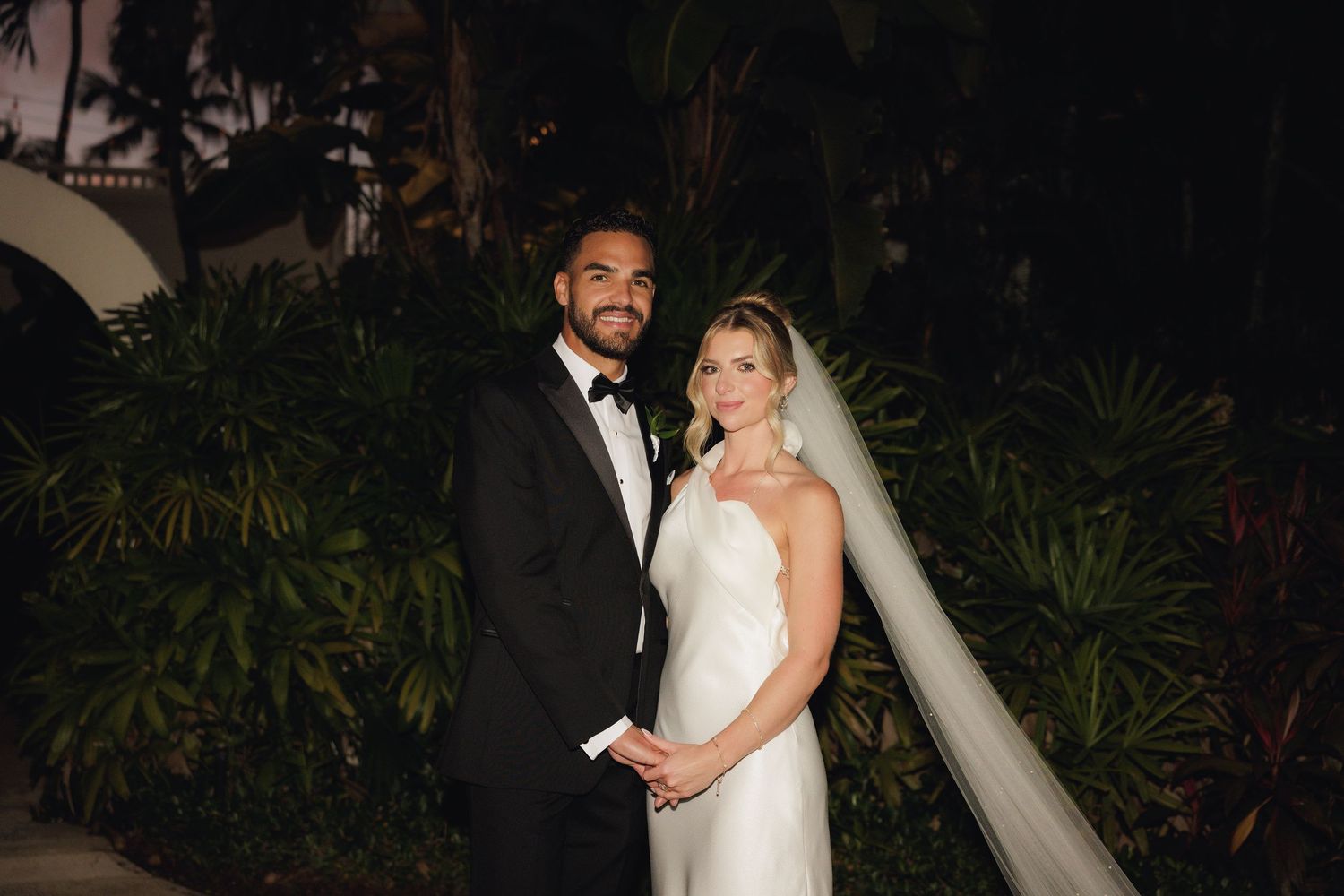A formally dressed couple in black tuxedo and white wedding dress pose together in an evening garden setting.