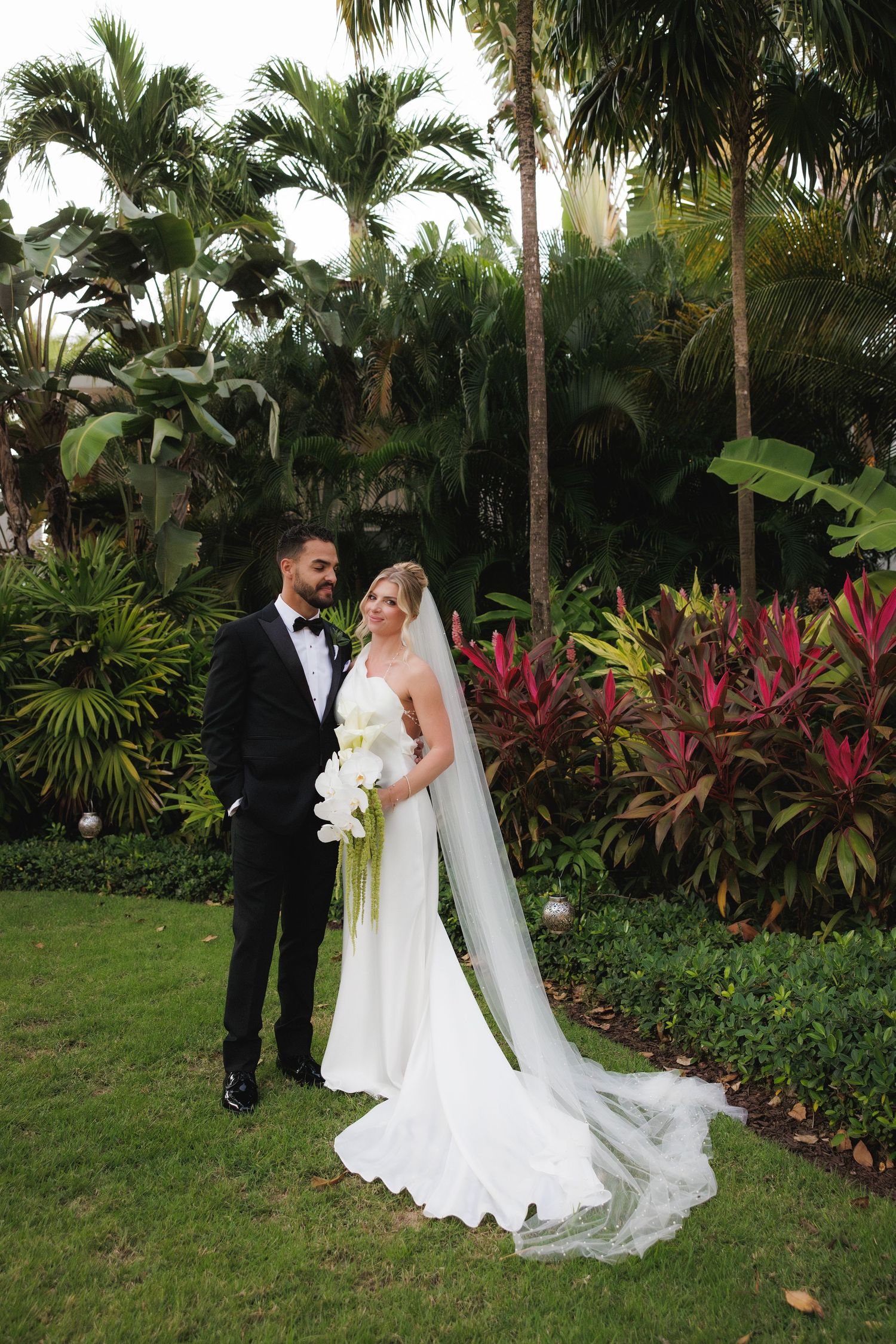 Newlyweds pose together on a tropical garden lawn surrounded by lush palm trees and vibrant red flowering plants.
