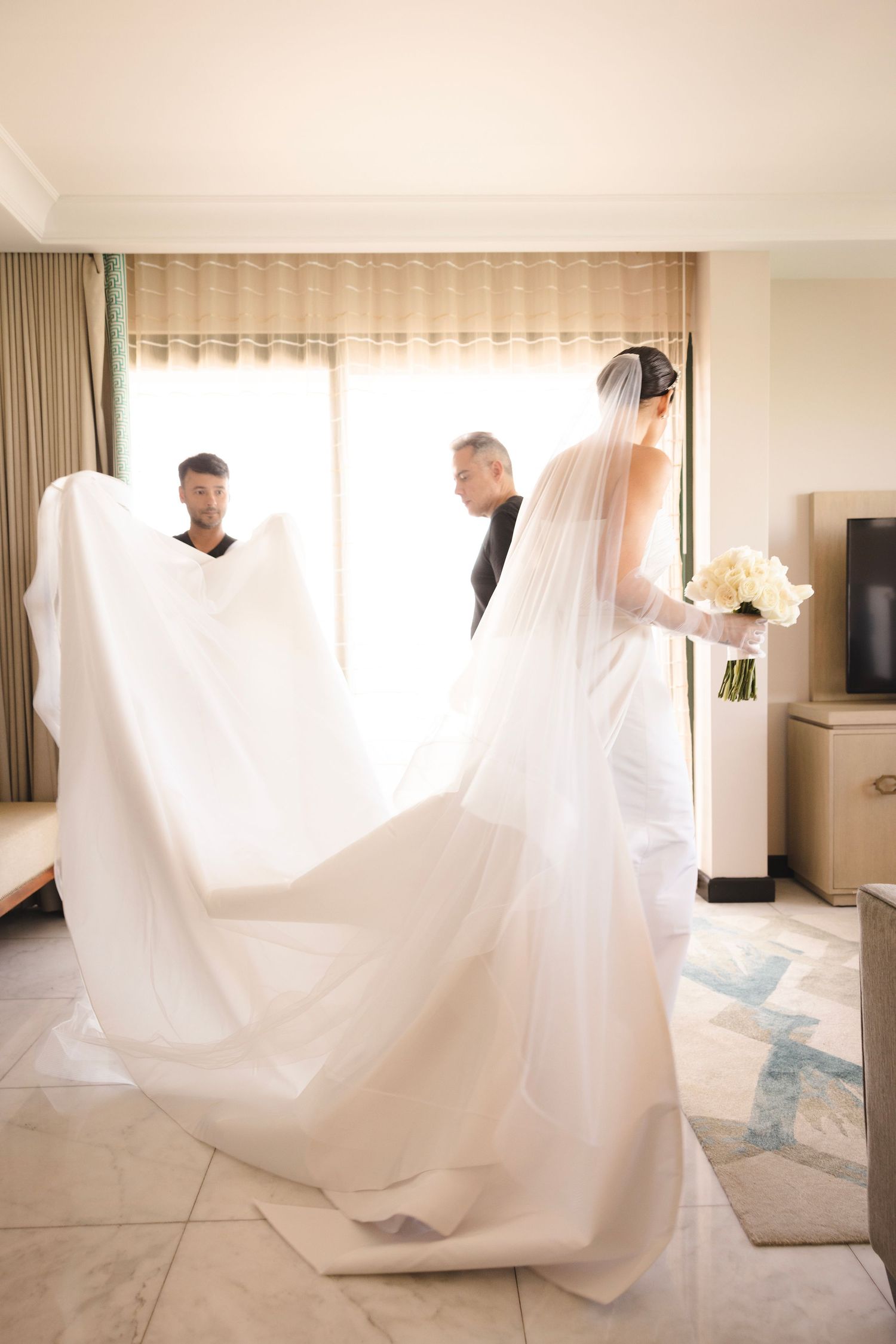Two people help adjust a flowing white wedding dress train and veil in an elegant hotel room interior.
