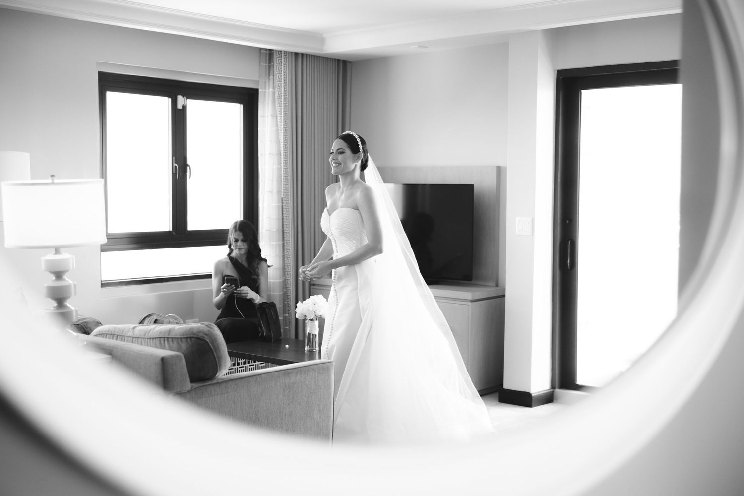 Black and white reflection through mirror showing elegant white wedding gown in a bright hotel room.