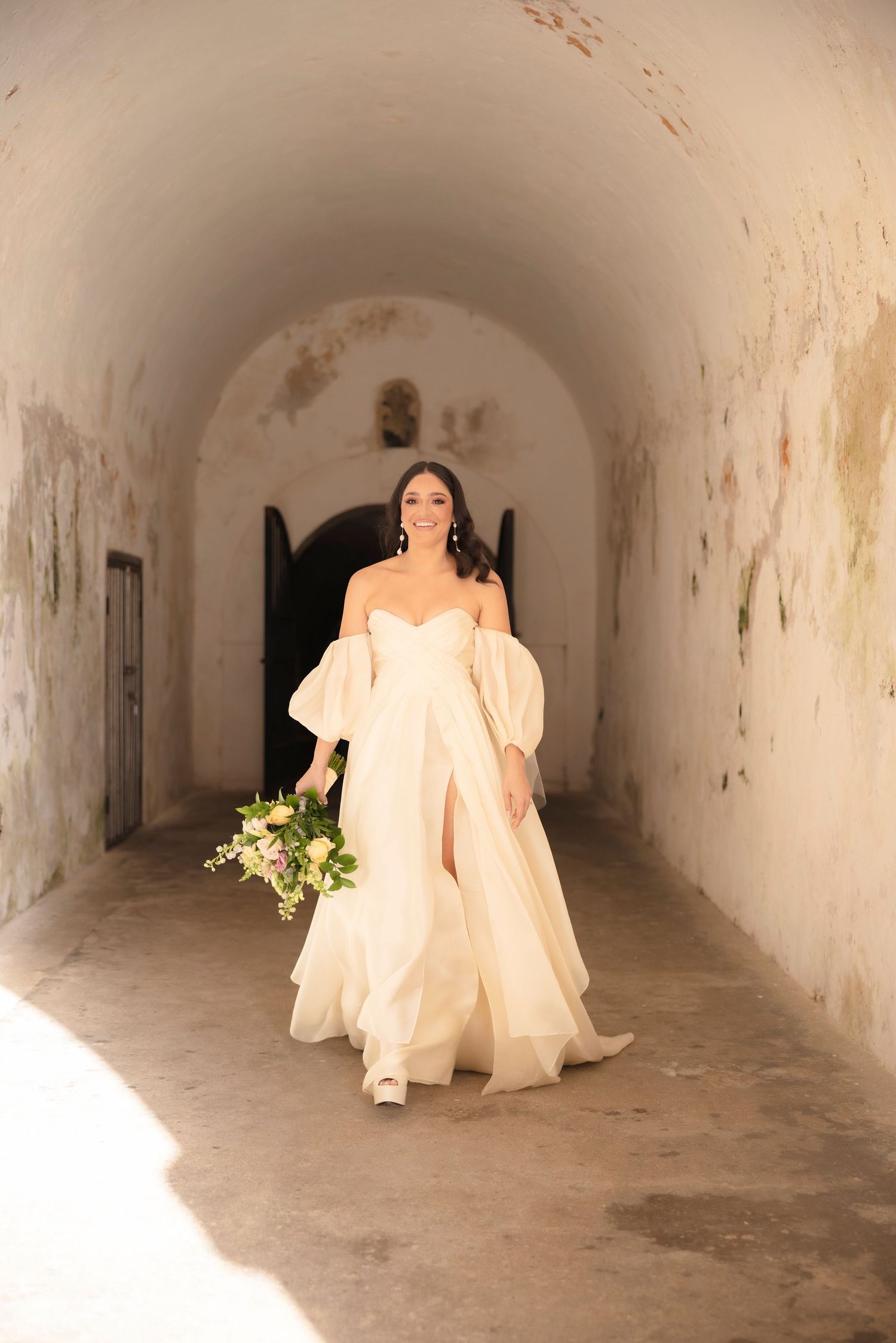 A flowing white wedding gown moves through a sunlit tunnel with textured stone walls.