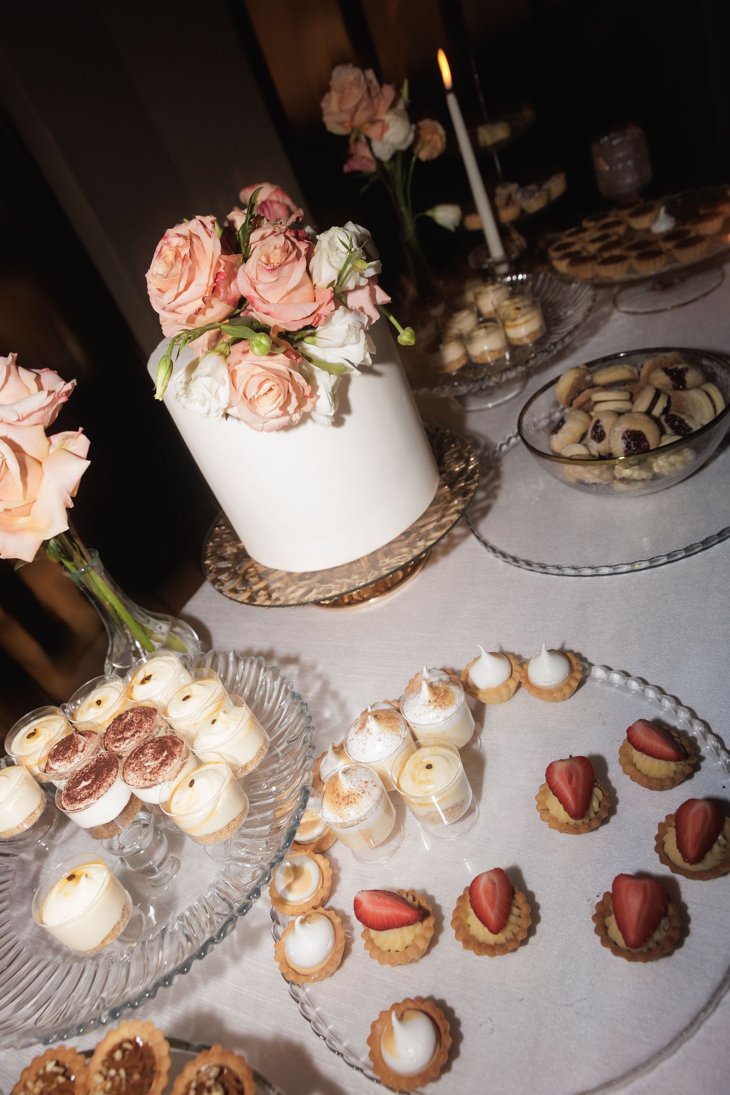 Elegant dessert table with tiered white wedding cake adorned with pink roses and small decorated pastries on glass platters.