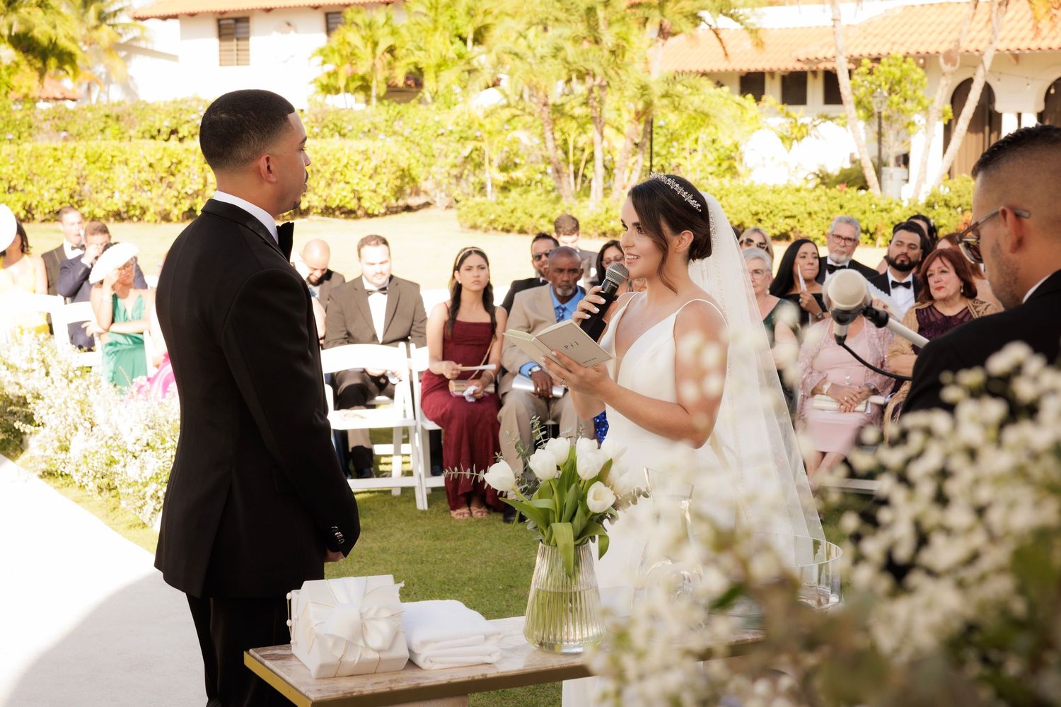 Couple exchanges wedding vows during outdoor garden ceremony surrounded by guests and white floral decorations.