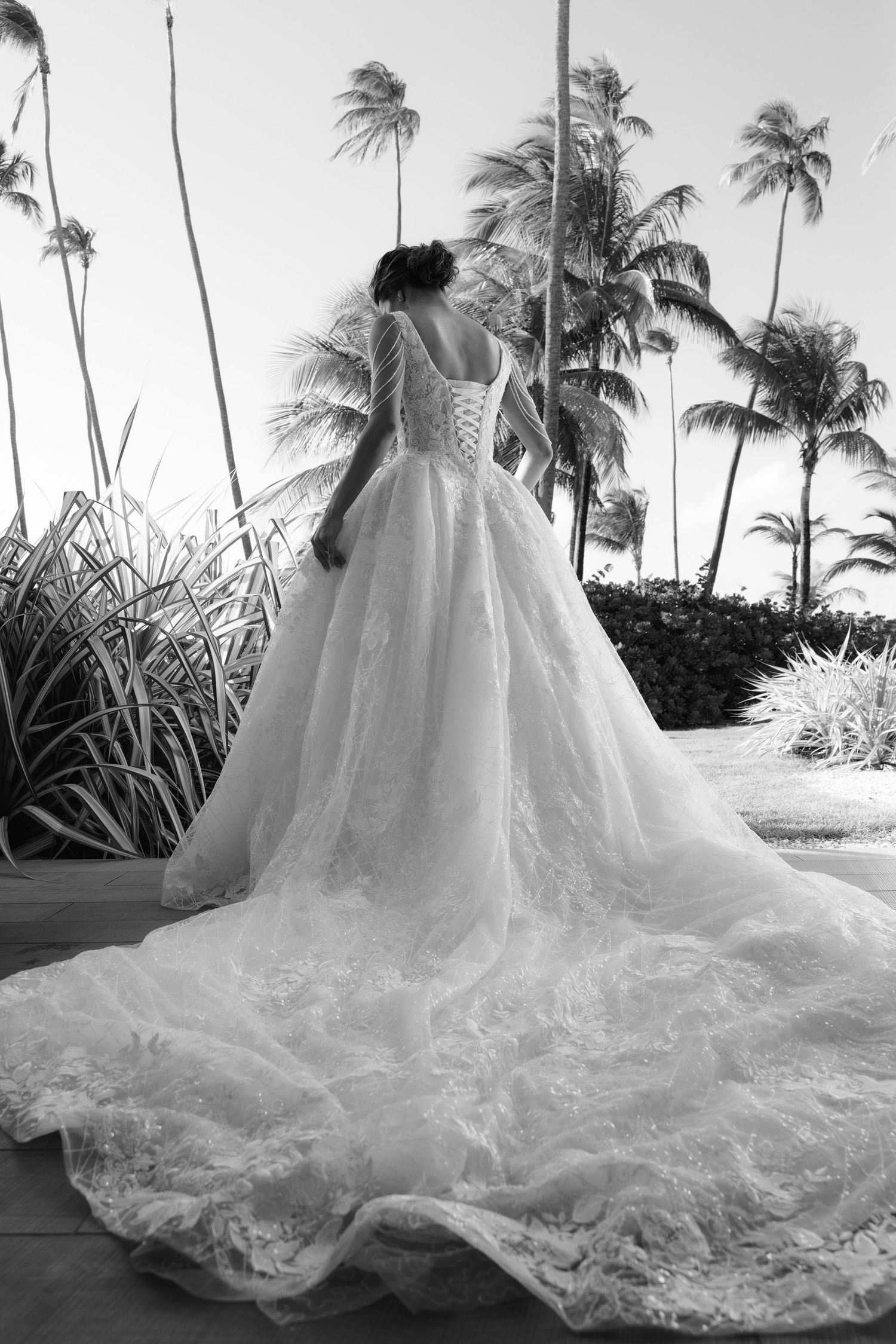 A flowing white wedding dress with a dramatic cathedral-length train photographed against tropical palm trees on a beach.