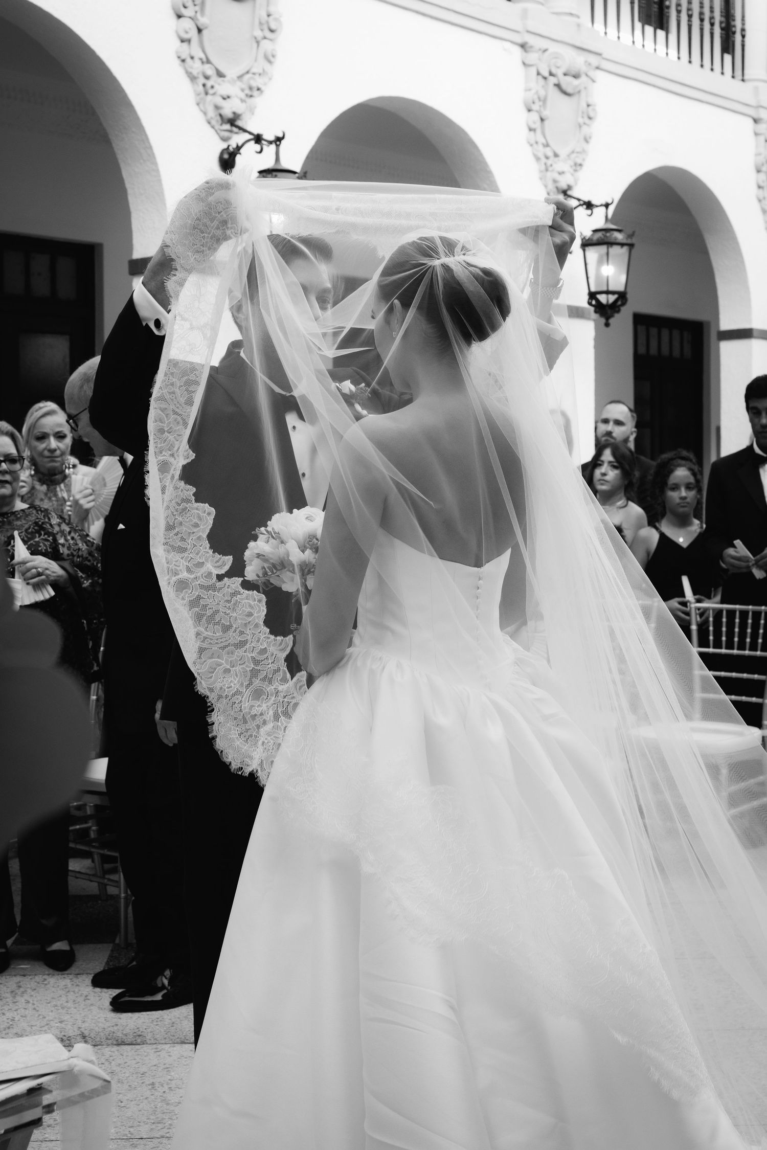 Black and white photo of a bride in a flowing veil surrounded by guests at an ornate outdoor venue.