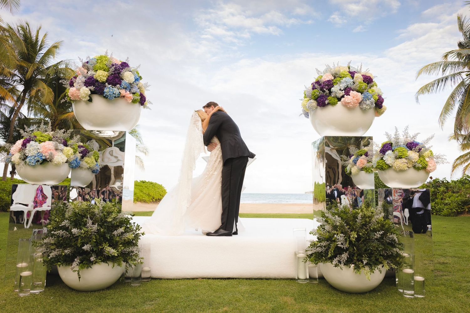 A beachside wedding ceremony setup with white floral arches and tropical palm trees in the background.