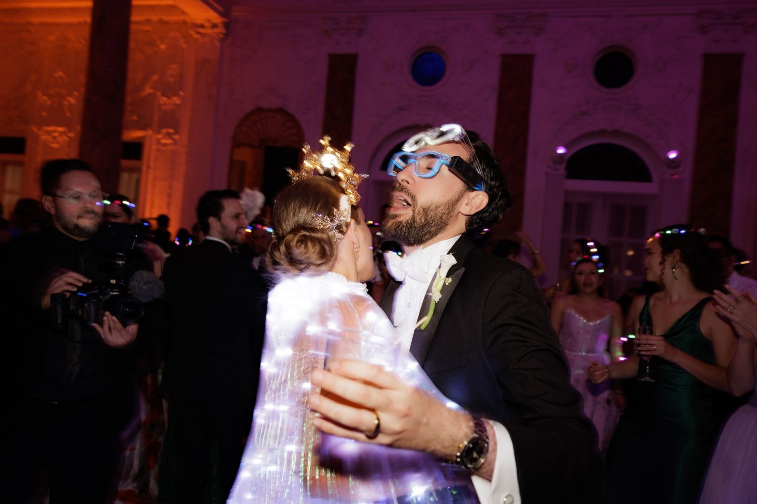 Dance floor filled with celebrants during a wedding reception under purple and amber lighting.