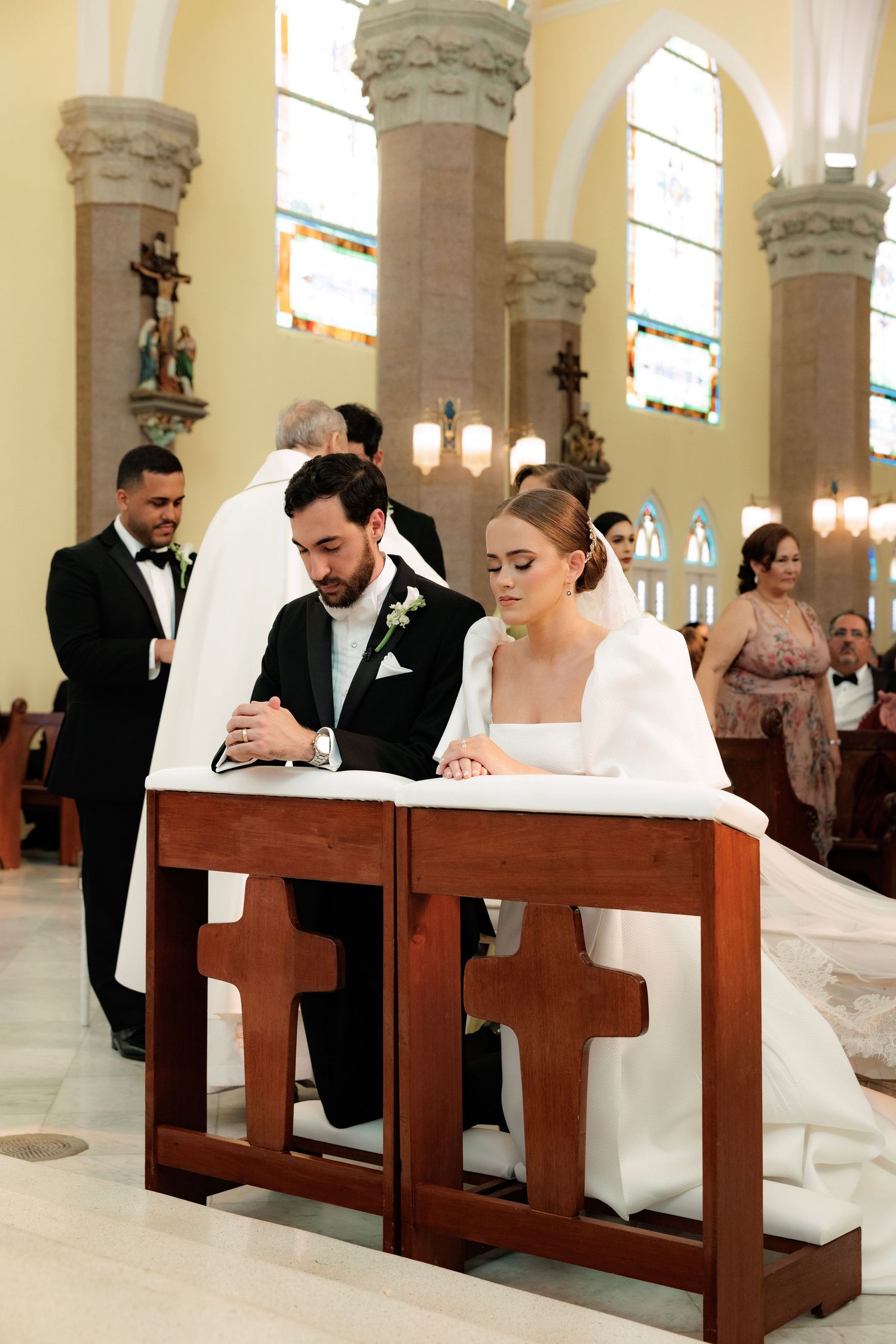 A couple signs documents while seated at wooden kneelers inside an ornate church with stained glass windows and columns.