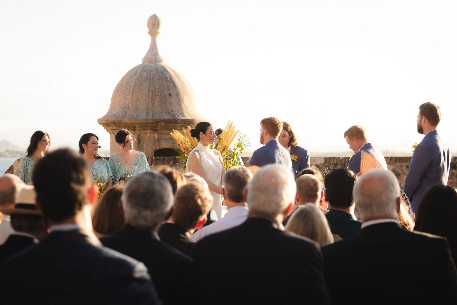 Wedding guests watching an outdoor ceremony with a domed structure visible in the background at sunset.