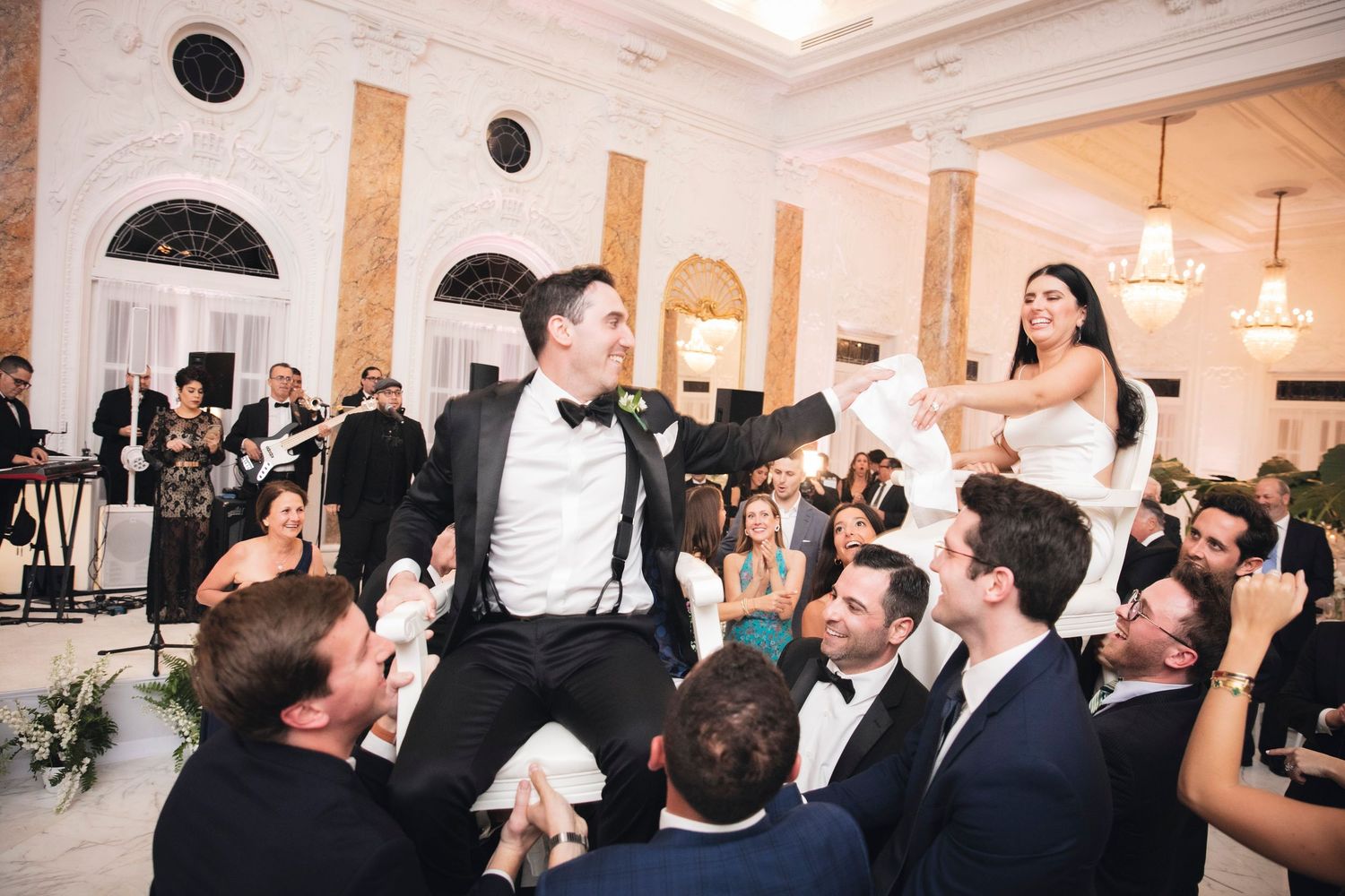 Wedding celebration scene showing guests lifting and celebrating in an elegant ballroom with classical architecture.