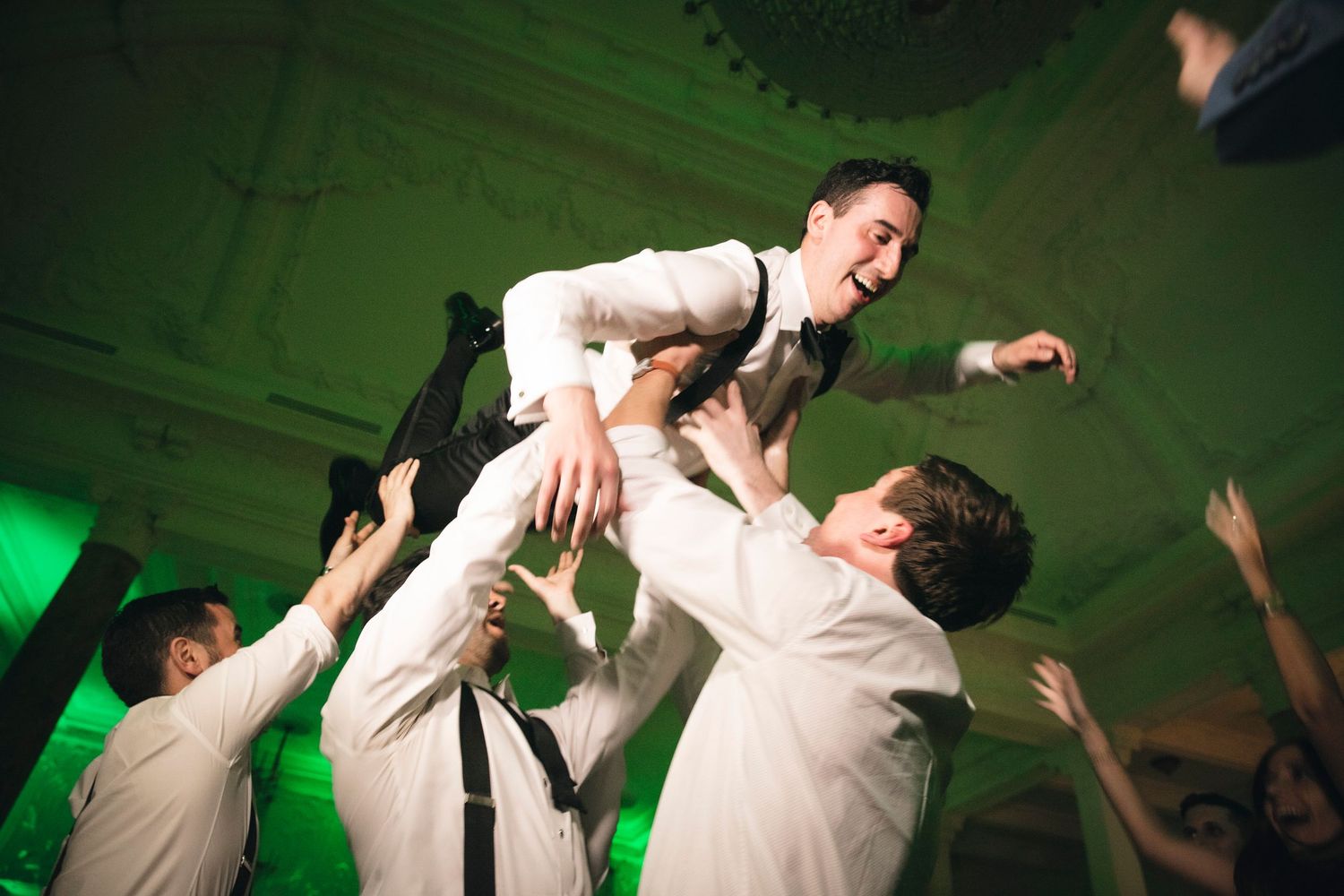 Wedding guests lift and celebrate with loved ones on the dance floor under green lighting at a reception party.