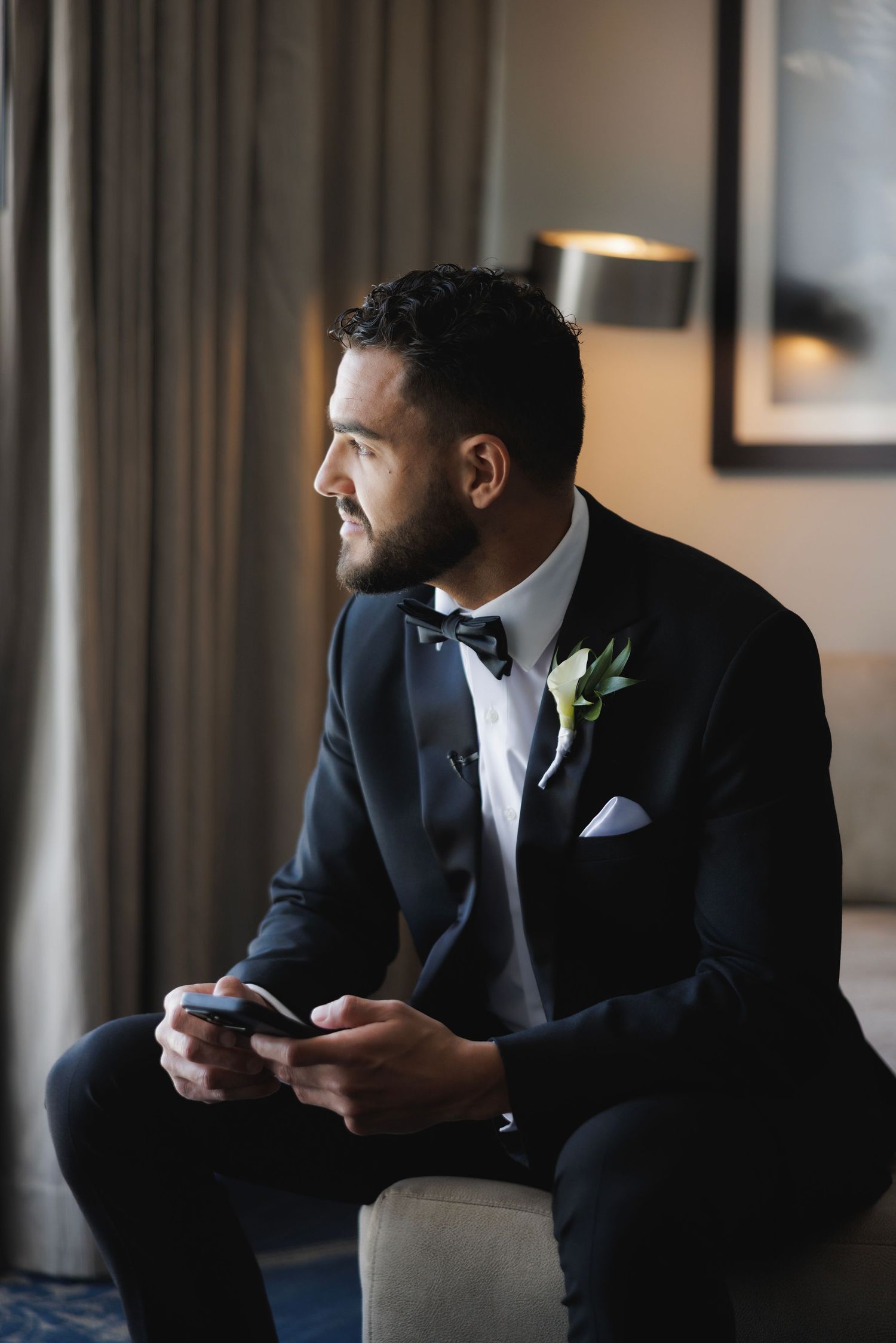 A groom in a classic black tuxedo and bowtie sits with phone in hand near a window in warm lighting.