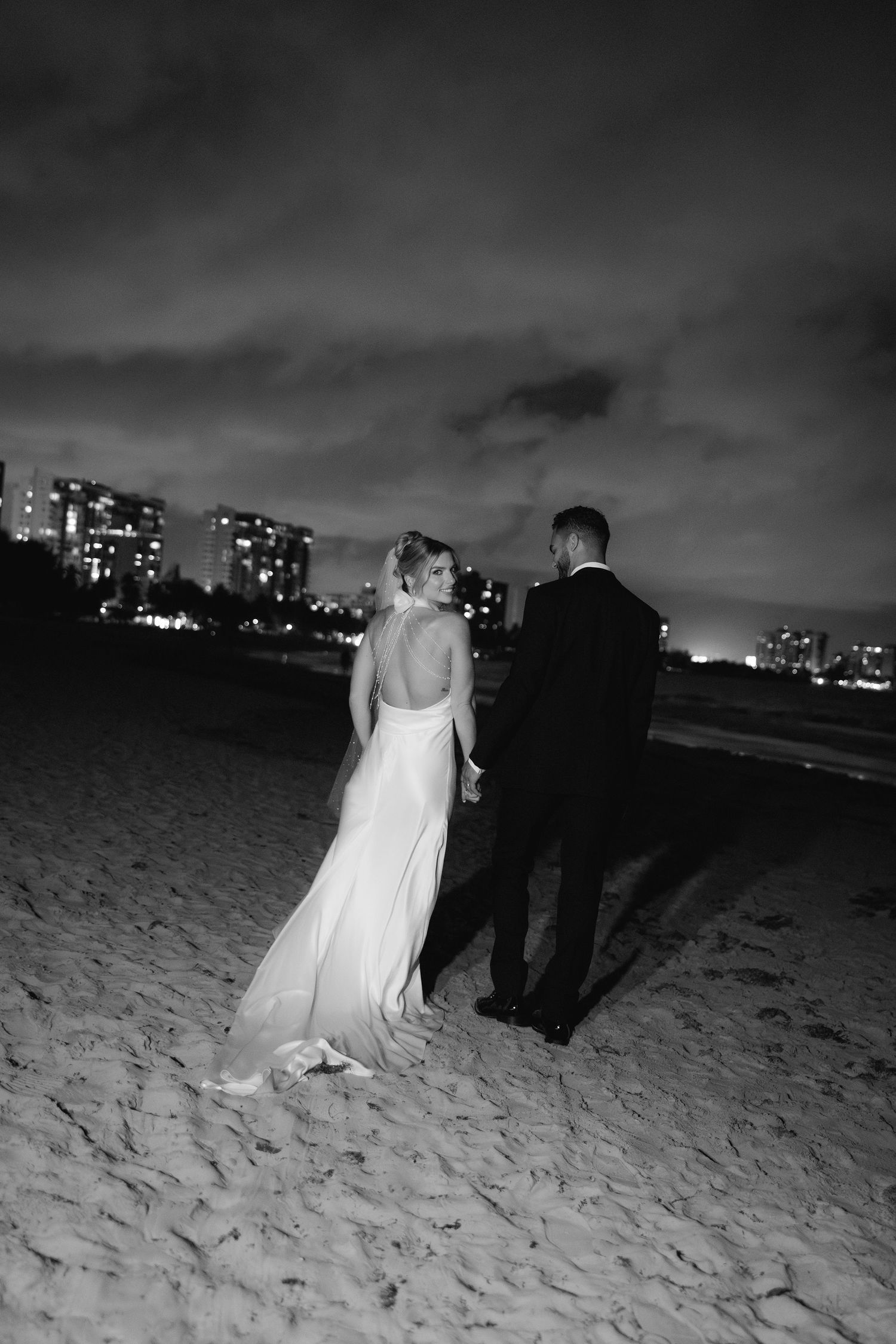 A couple in wedding attire walks along a dark beach at night with city lights glowing in the distance.