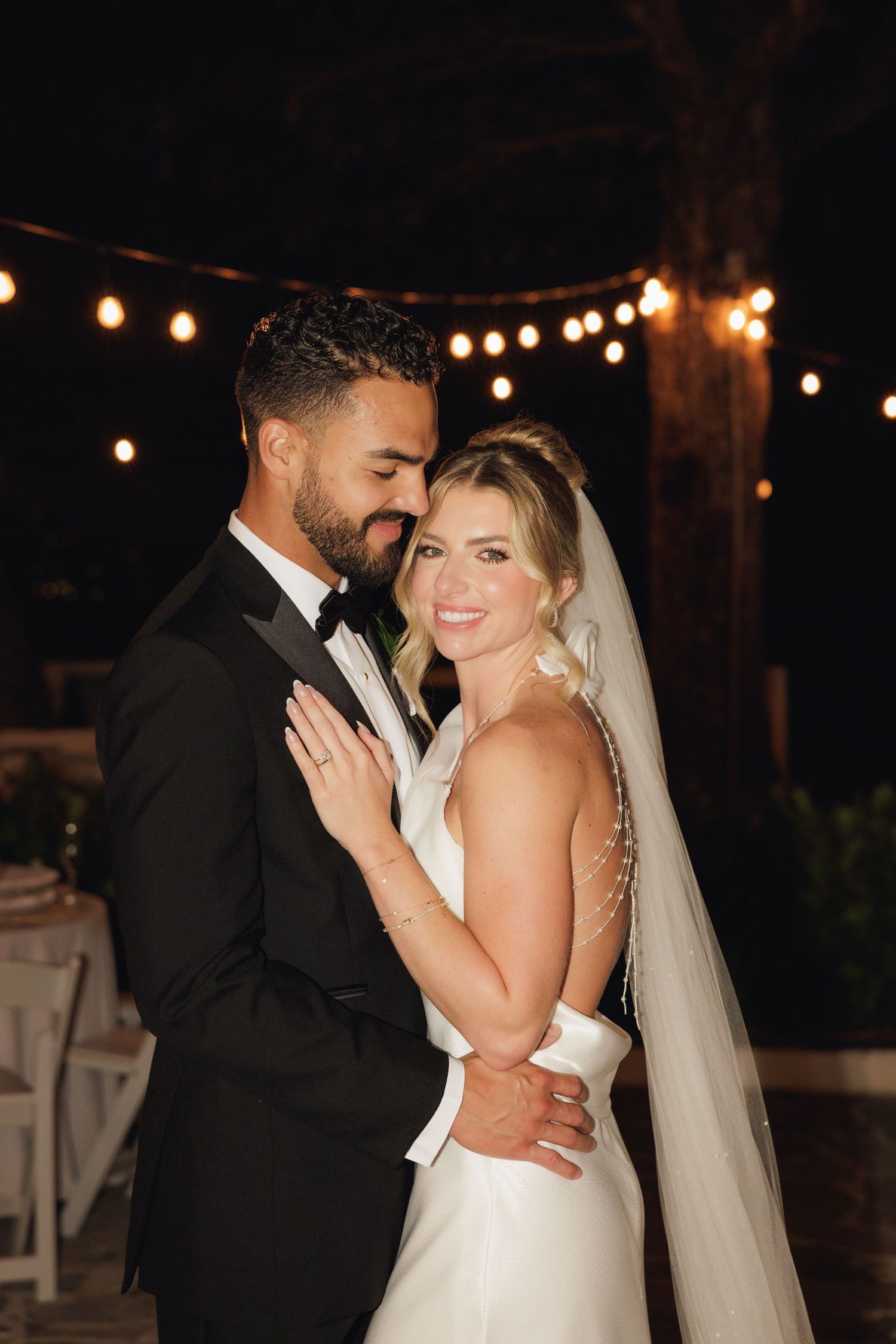 A newlywed couple shares an intimate moment under string lights at their evening outdoor wedding reception.