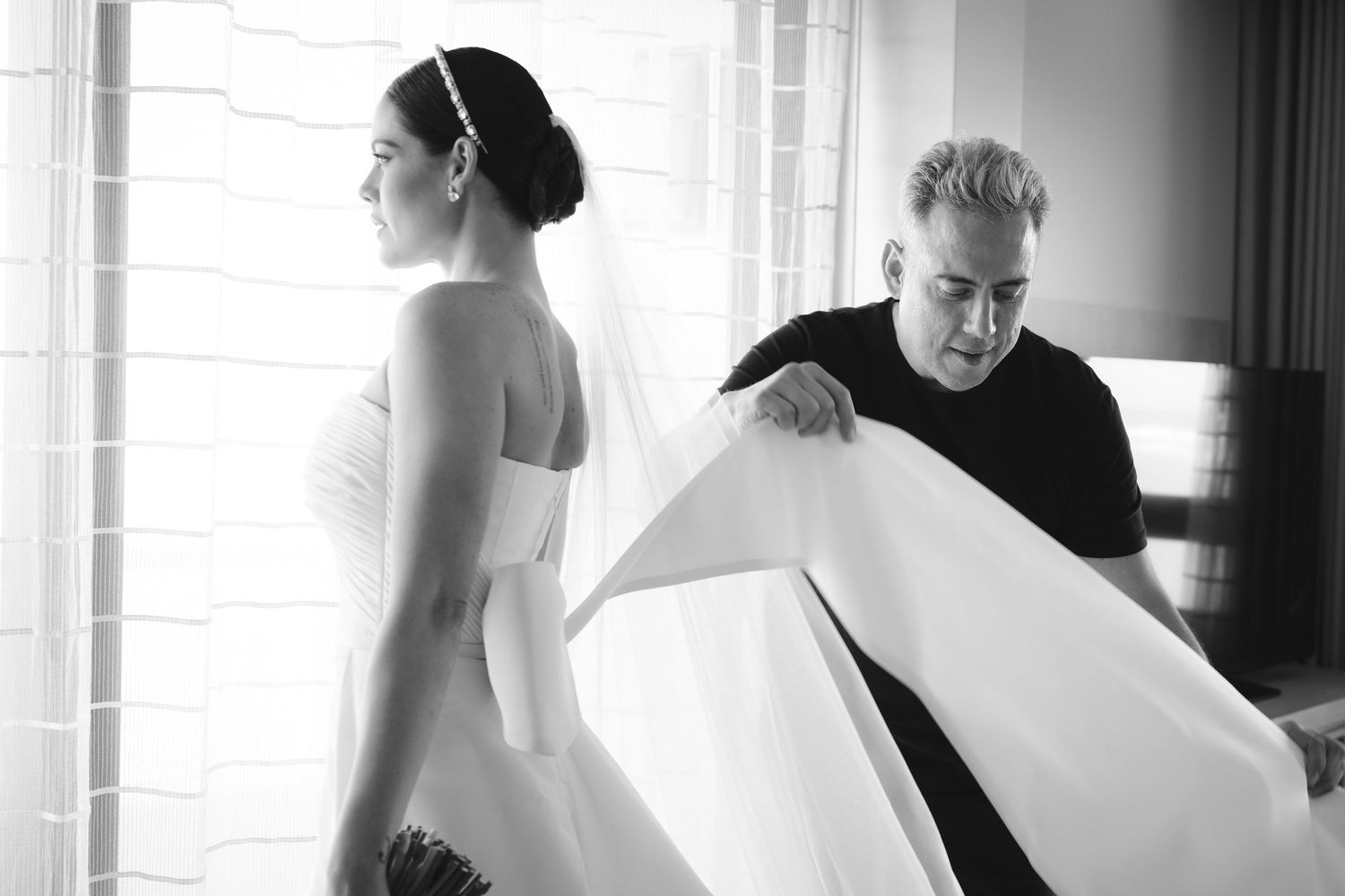 A black and white photograph shows a bride in a wedding gown getting final adjustments on her dress.