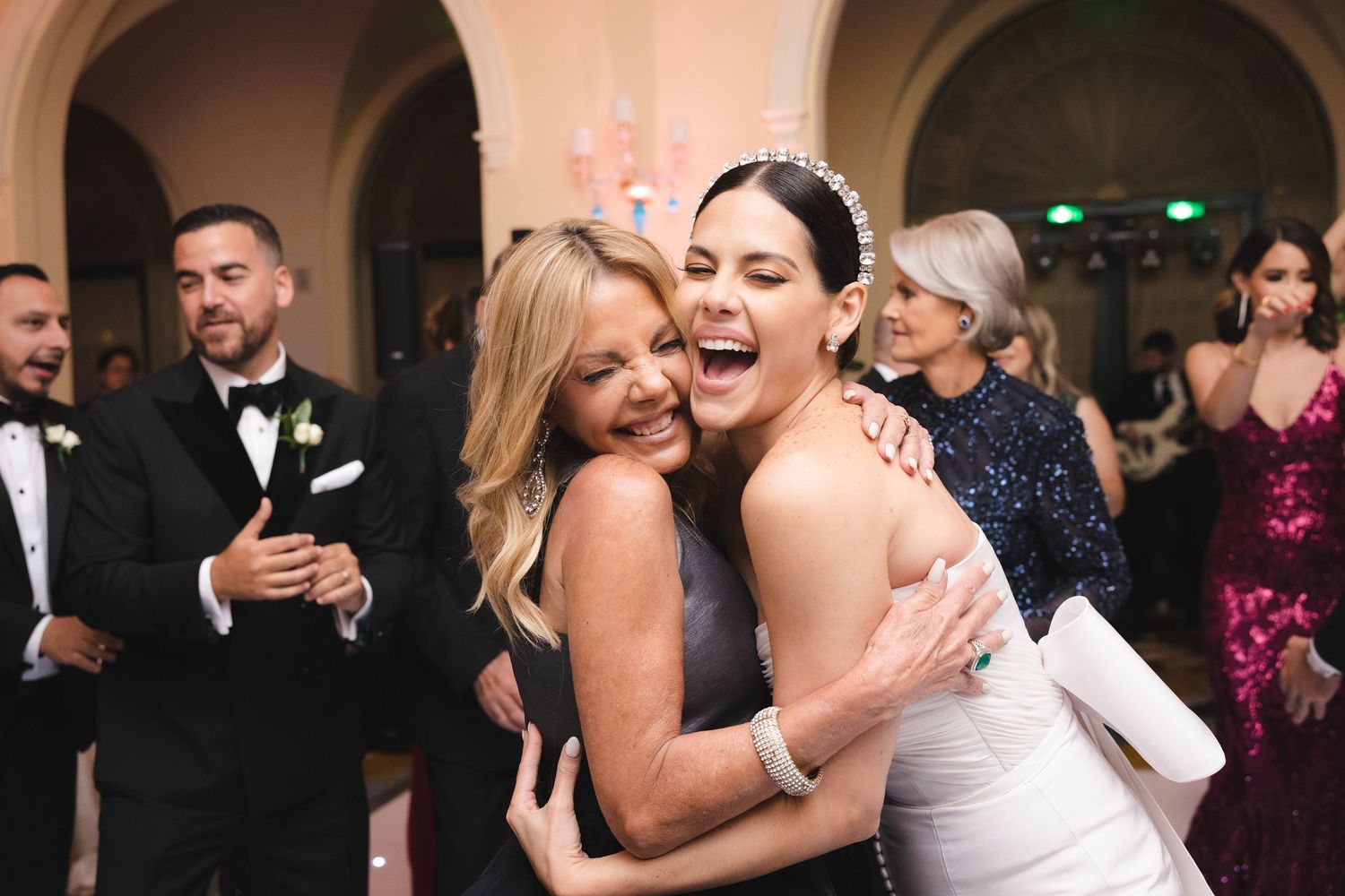 Two joyful people share a candid laughing moment during a formal wedding reception with guests in background.
